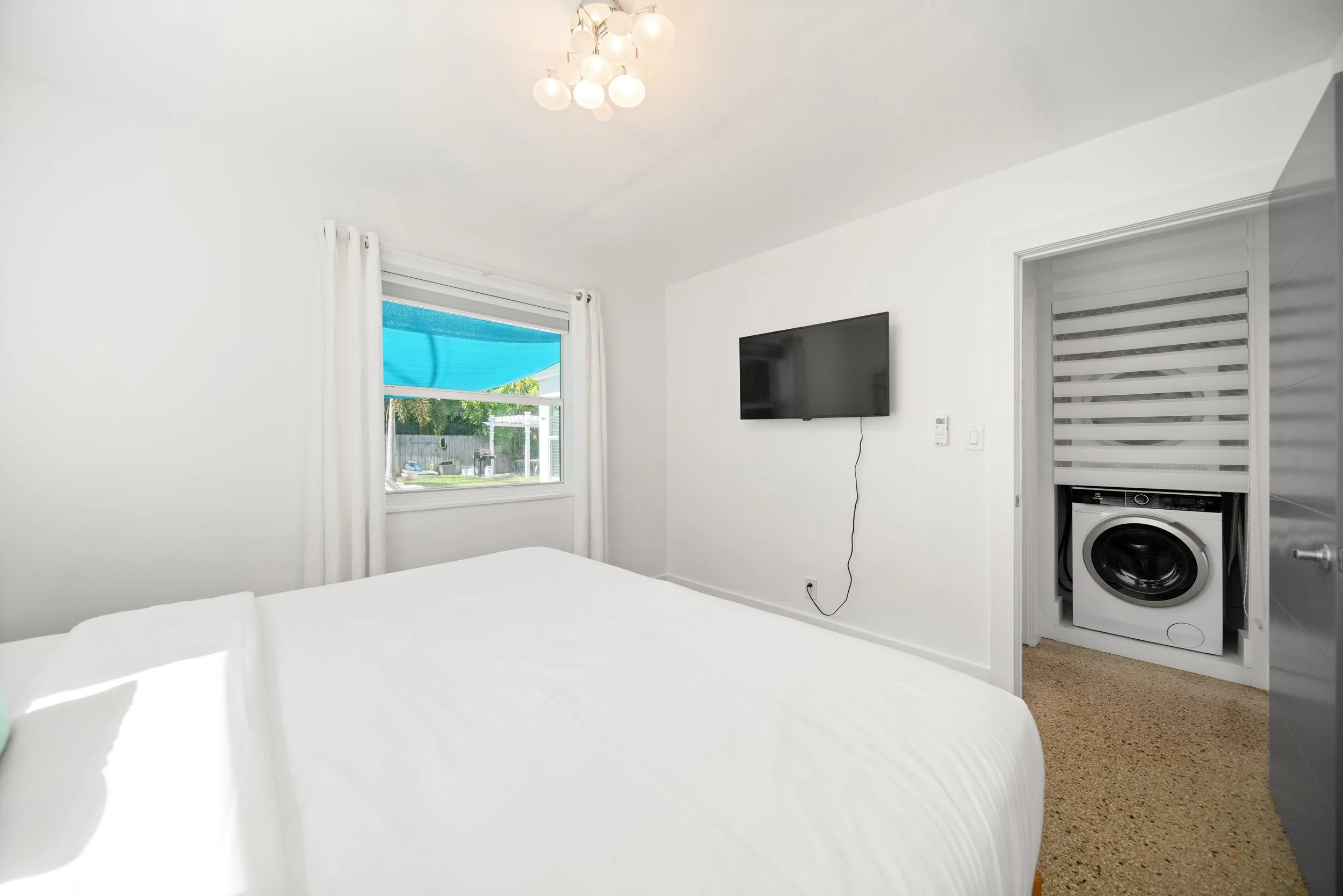 A white bedroom with a bed, a window with white curtains, a wall-mounted TV, and an open closet with a washing machine inside.