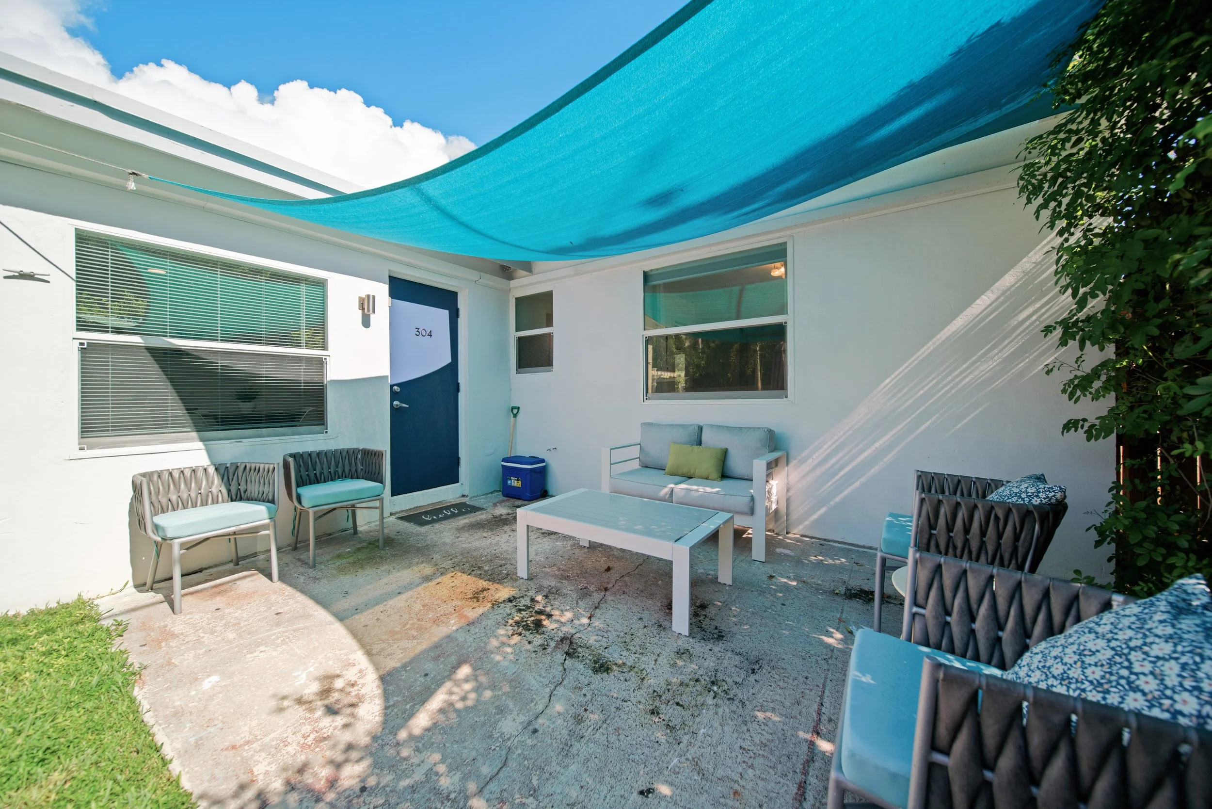 An outdoor patio area with outdoor furniture including chairs with cushions and a table, a white bench, a sofa, a blue door with the number 304, and a blue shade cloth overhead, with shadows and greenery visible.