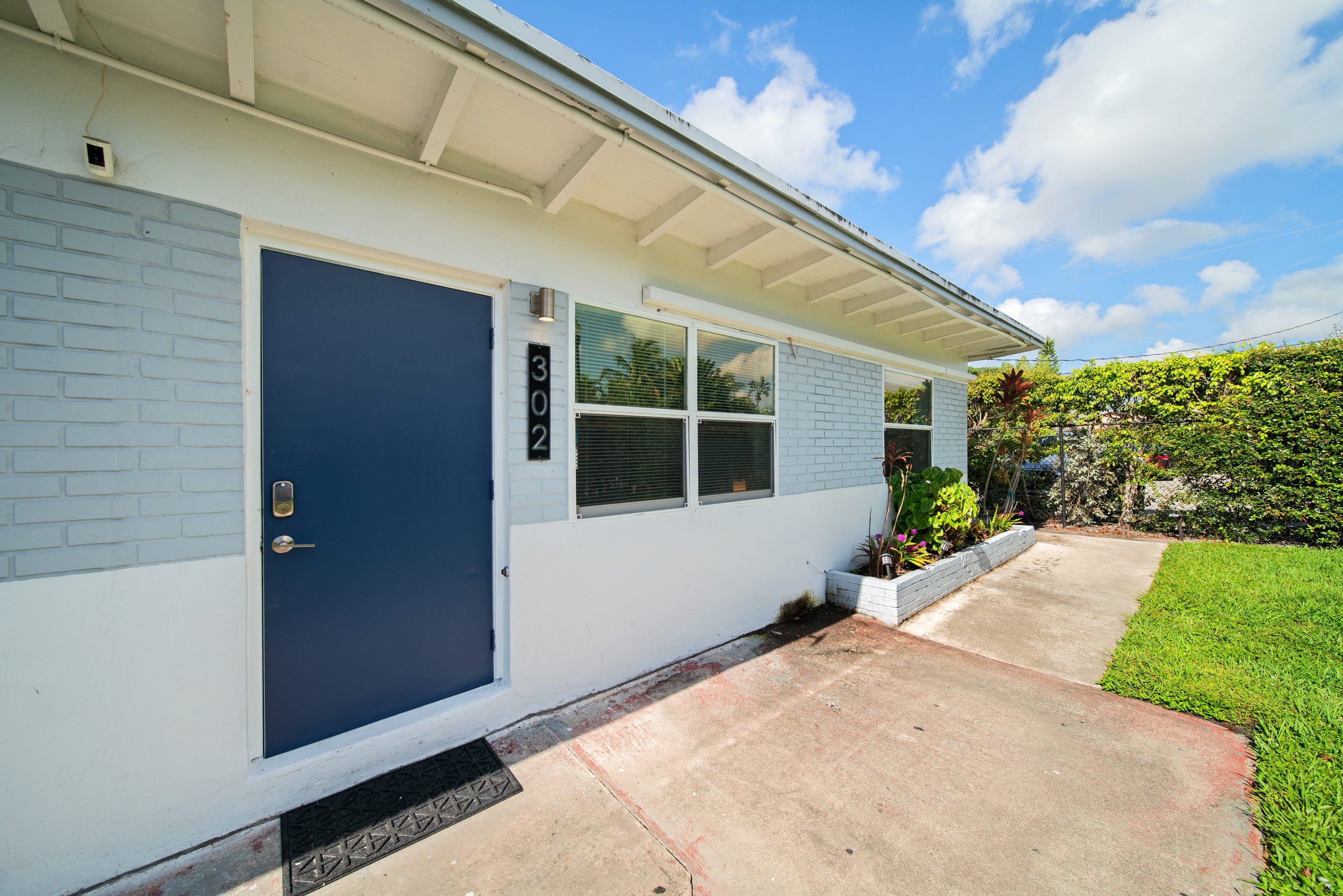 Front view of a modern house with a blue door, white brick walls, and two large windows. The house has a white overhanging roof, a small garden with plants along the front, and a concrete sidewalk leading to the door. The sky is partly cloudy with bl