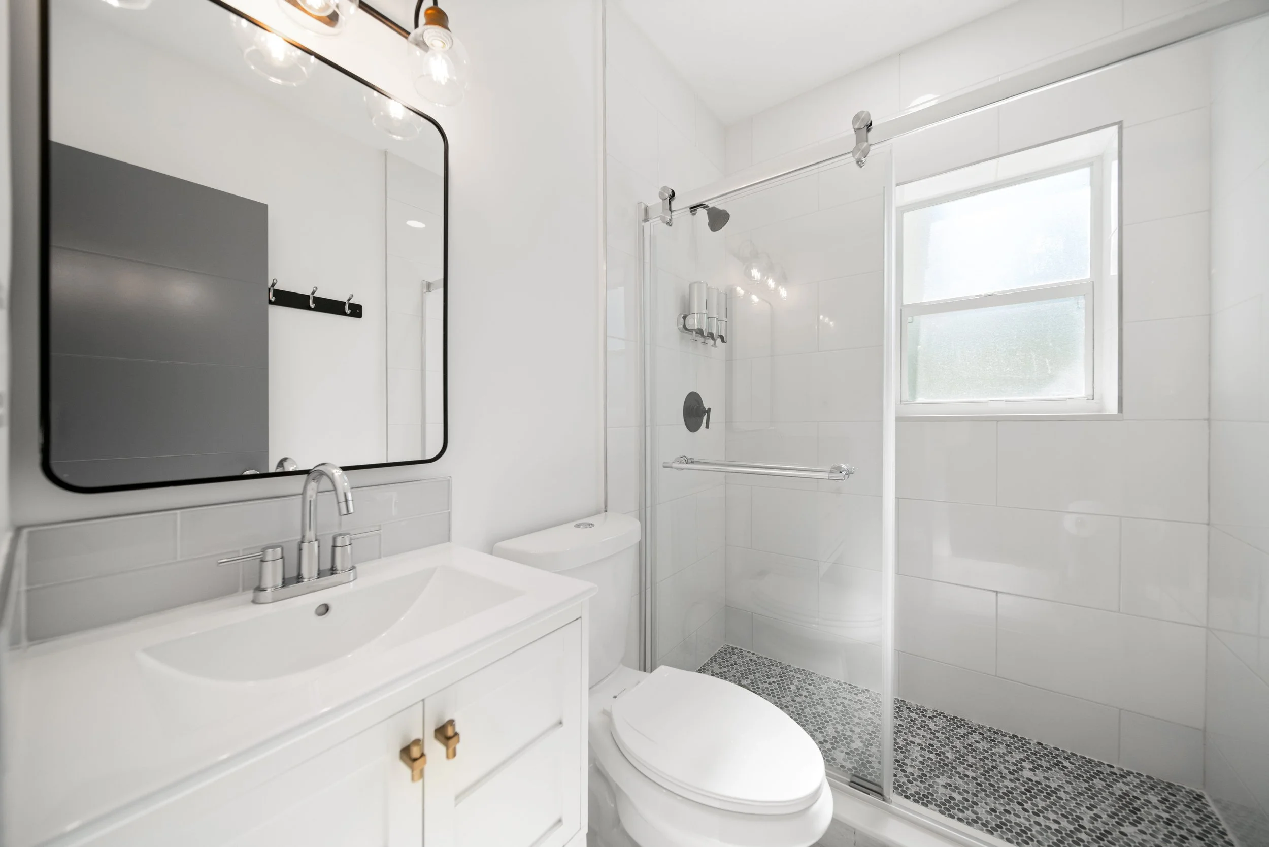 White bathroom with a sink and vanity, a toilet, a shower with glass door, gray tiled shower floor, and a window.