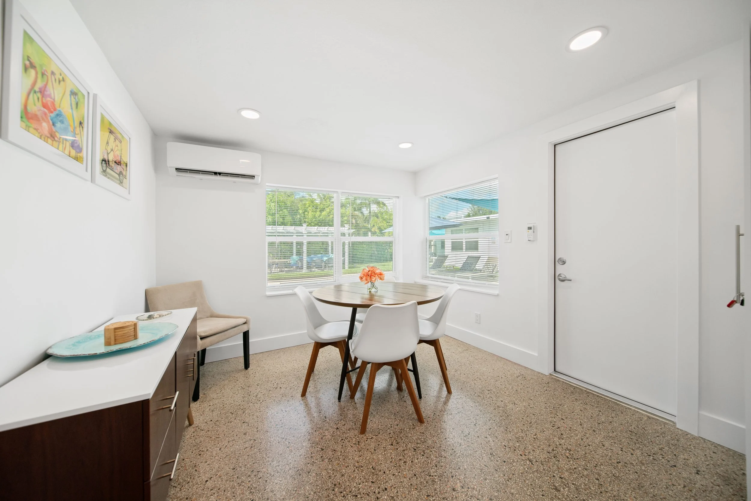 A bright, modern dining area with white walls and large windows showing a green outdoor landscape. There is a round wooden table with white chairs, a beige armchair, and a cabinet with decorative items. Artworks hang on the wall, and recessed lightin