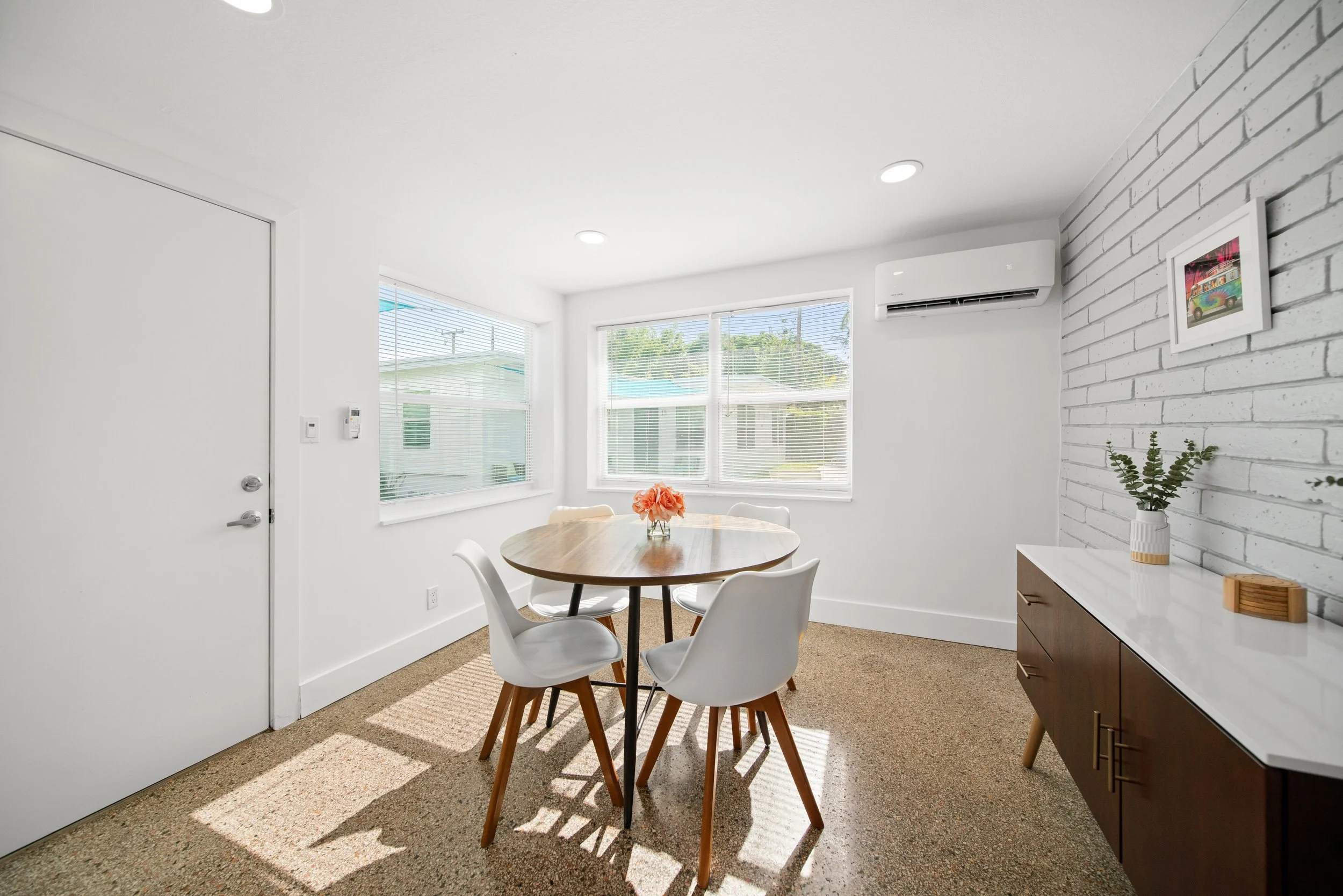 A bright dining area with large windows, a round wooden table, four white chairs, a decorative flower vase, a white and brown sideboard with a plant on top, and a white brick accent wall with a colorful picture frame.

