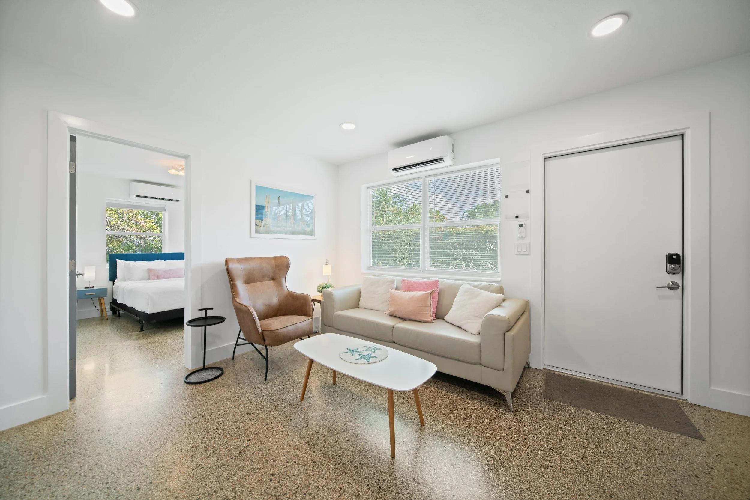 Living room with beige carpet, white walls, a cream-colored sofa with pink and white pillows, a brown leather armchair, a white coffee table with starfish decorations, large window with blinds, and artwork of a beach scene; adjacent bedroom with bed,