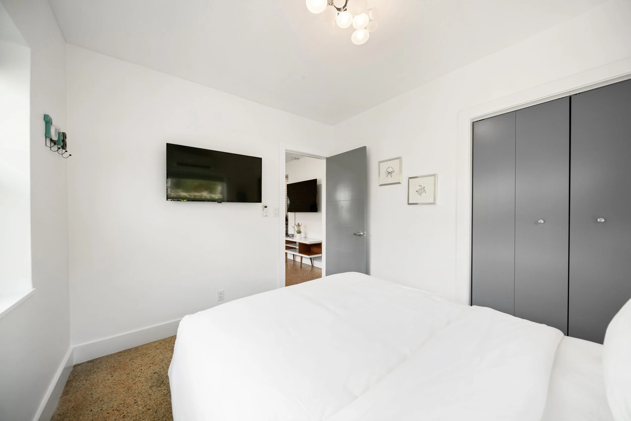 A clean, modern bedroom with white walls, a flat-screen TV mounted on the wall, a door, framed pictures, a gray closet, and part of a bed with white bedding.