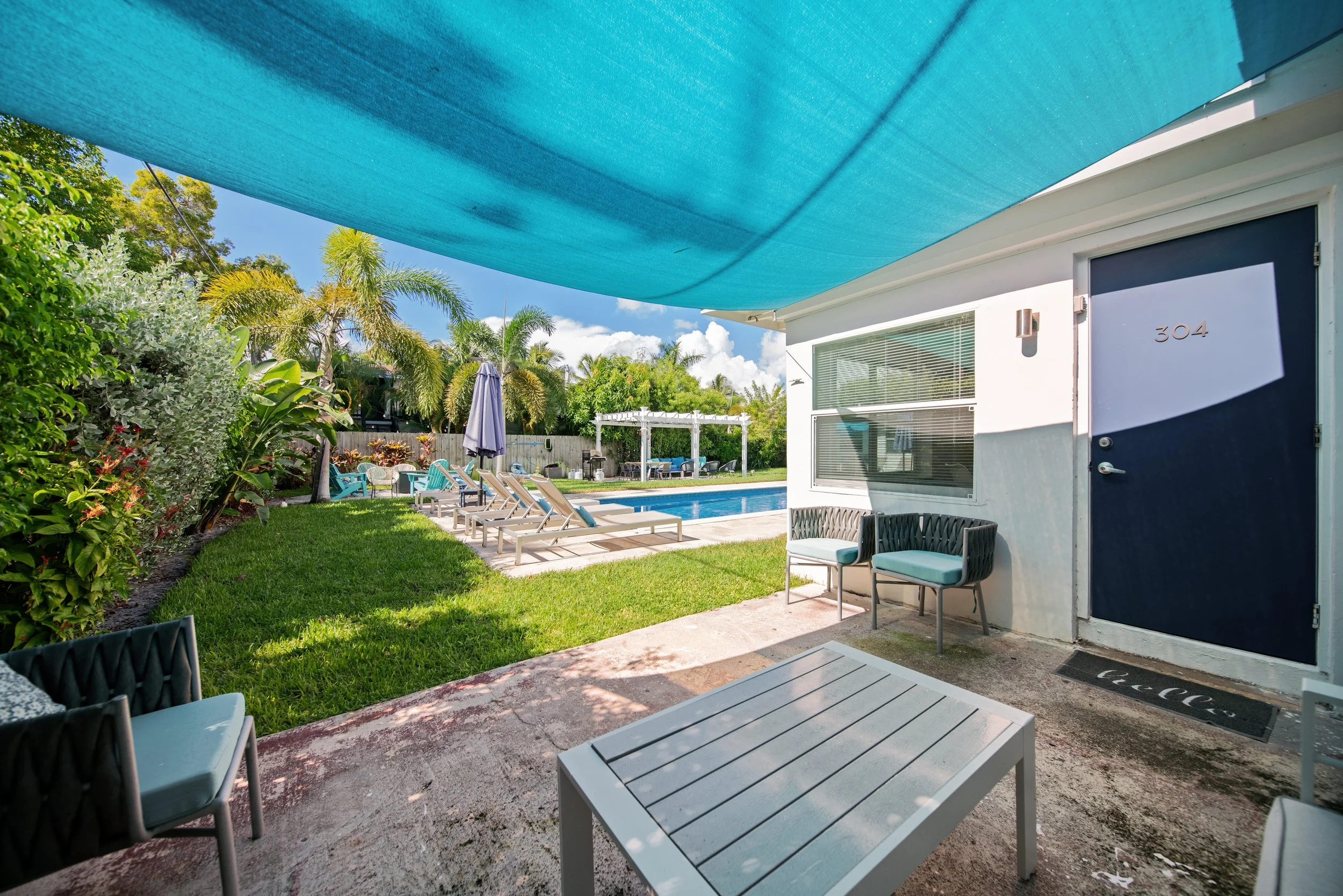 A backyard patio with a blue shade cloth overhead, surrounded by lush green plants and trees. There are several lounge chairs near a swimming pool, and a white pergola with outdoor furniture in the background. The house has a white exterior with larg
