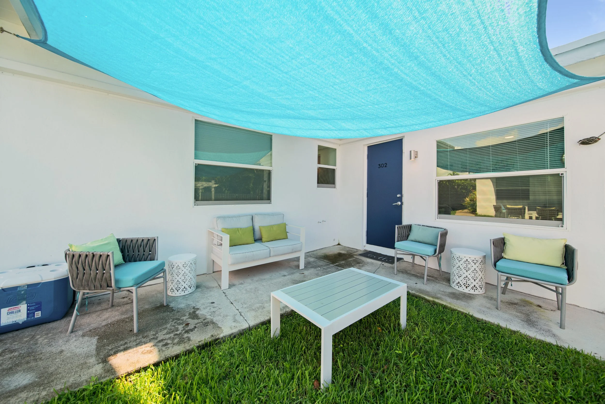 A covered outdoor patio with white walls and several pieces of outdoor furniture, including chairs with cushions, a bench, and a coffee table, under a blue fabric sunshade.
