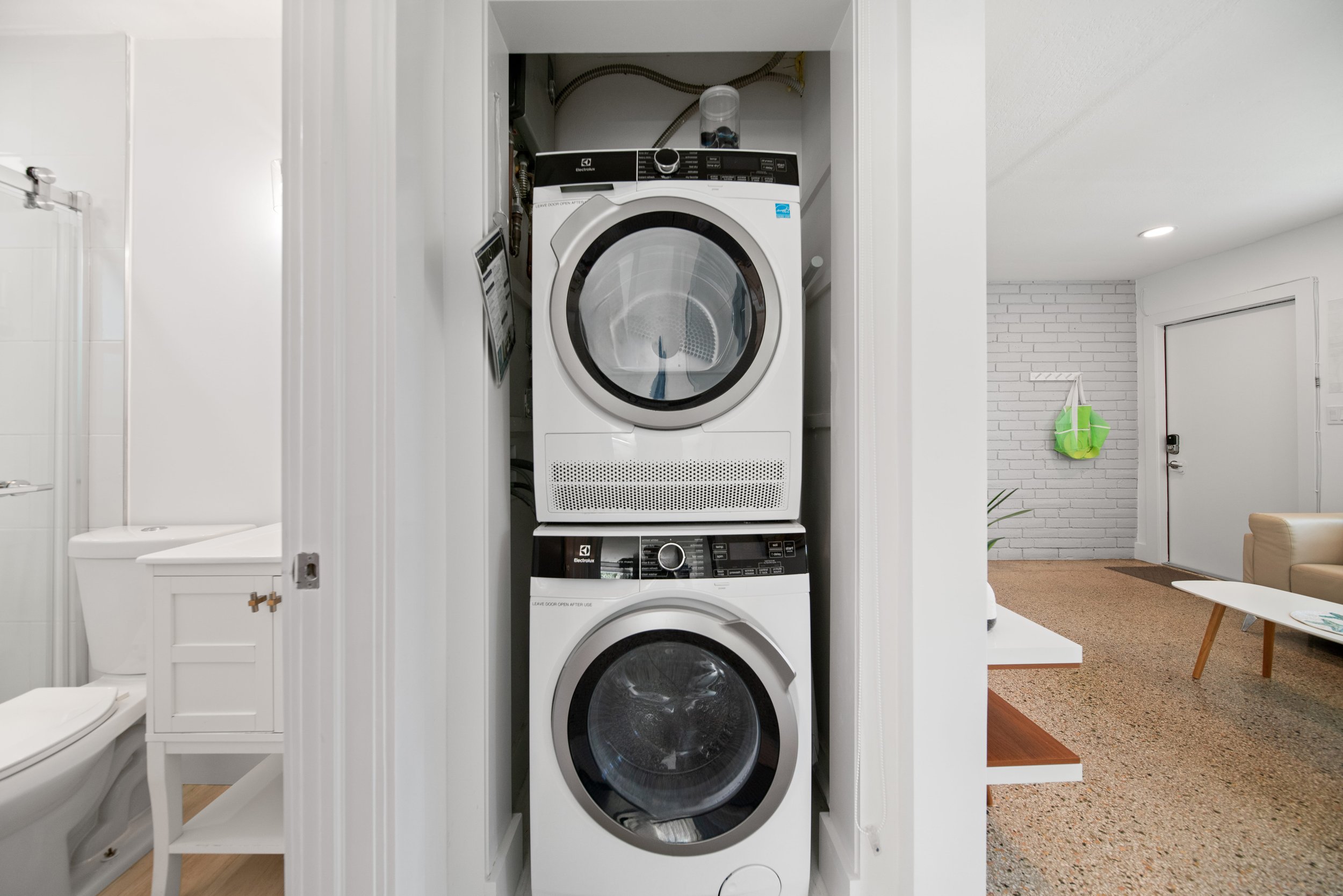 Stacked washer and dryer in a laundry closet with white trim, next to a partial view of a bathroom with a toilet and a white cabinet.