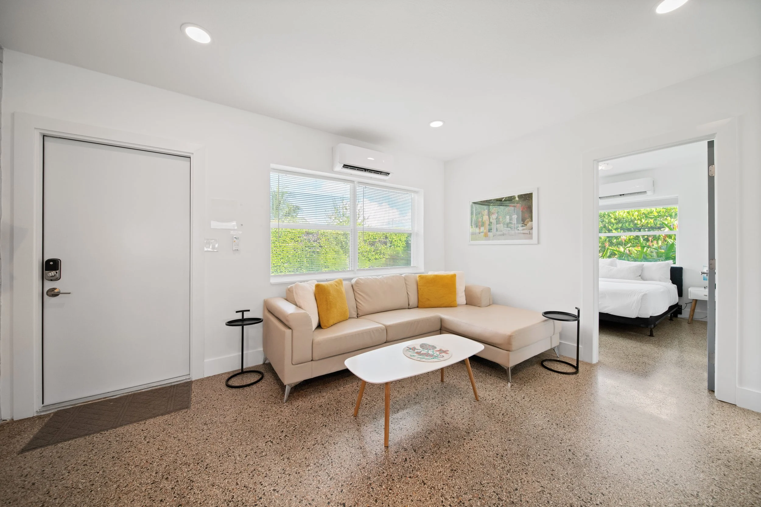 Living room with beige sofa, yellow cushions, small white coffee table with floral design, black side tables, large window with blinds, white walls, and bedroom visible through an open door.