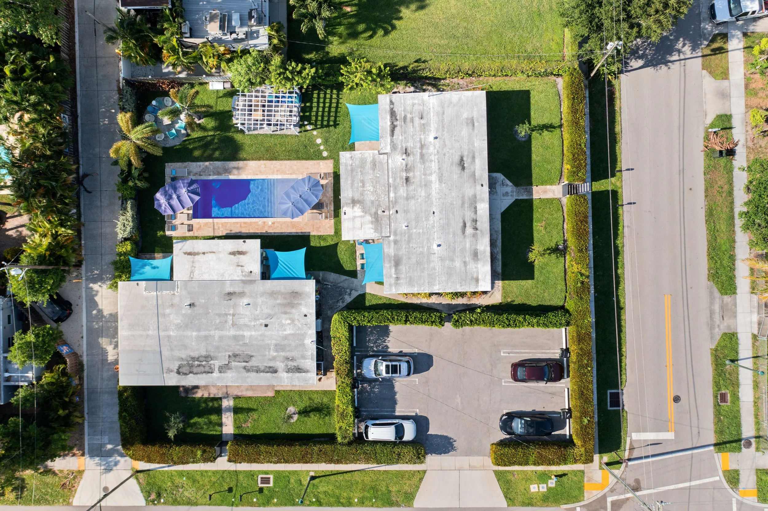 An aerial view of a residential backyard with a swimming pool, surrounded by greenery, trees, and outdoor furniture, adjacent to a parking lot and a street.