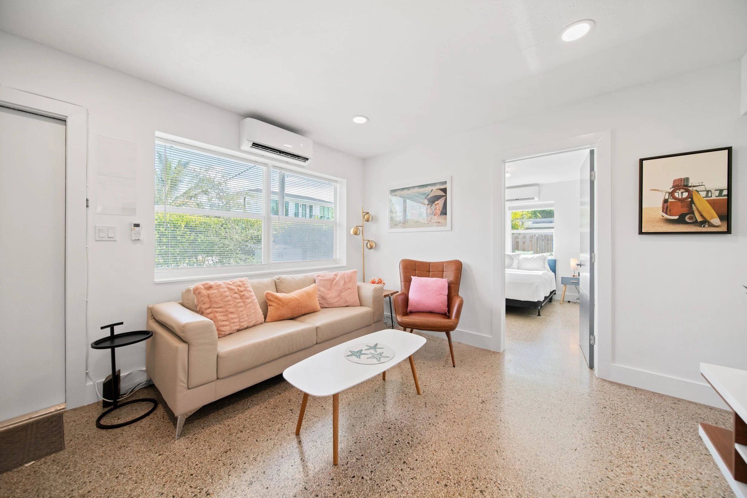 Living room with a beige sofa adorned with pink and beige pillows, a small white coffee table with starfish design, a brown armchair, and framed artwork on white walls. Large windows with blinds and an air conditioning unit are visible, with a bedroom seen through an open doorway.