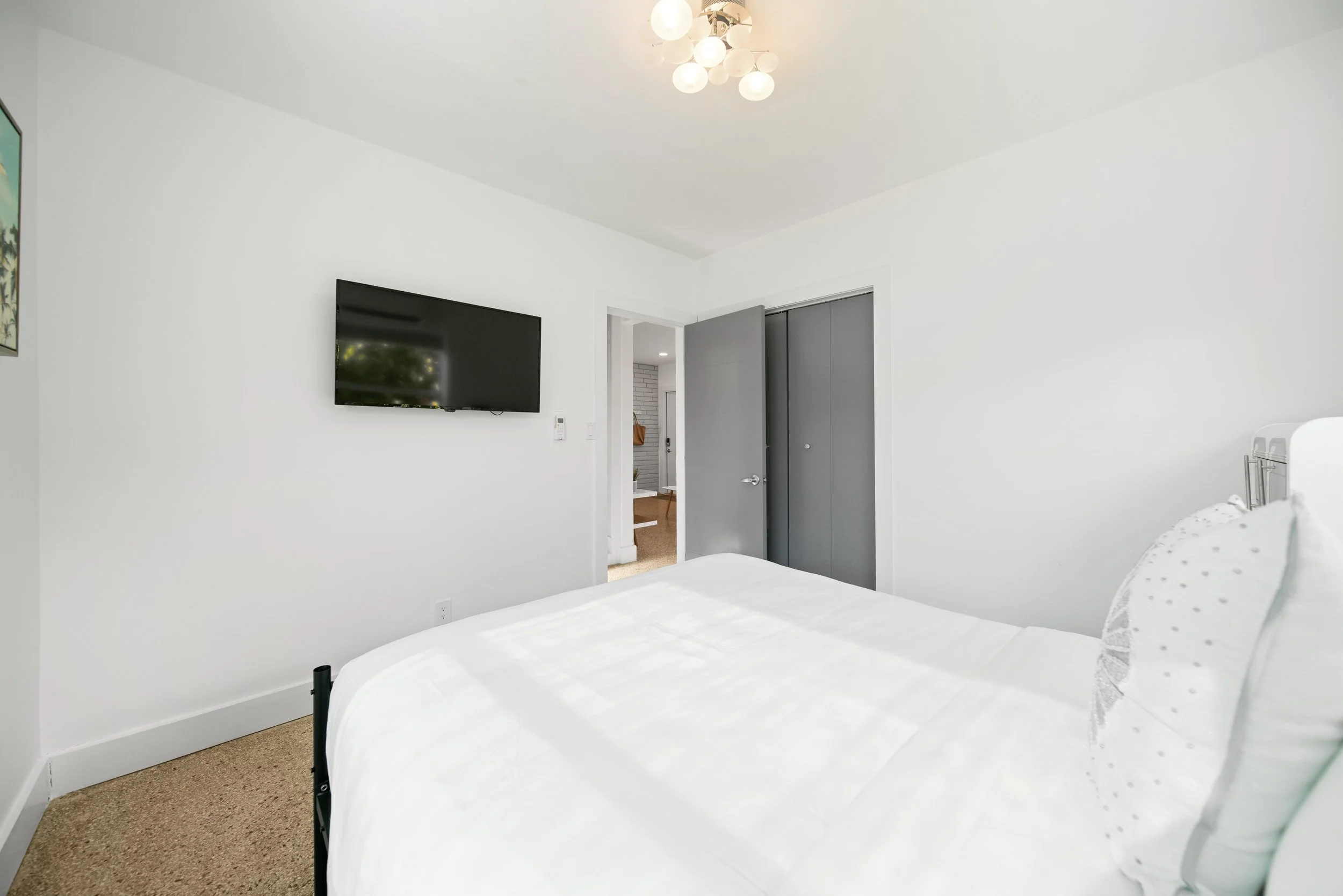 A bedroom with white walls and ceiling, a flat screen TV mounted on the wall, a door and closet, a bed with white bedding, and a modern ceiling light fixture.
