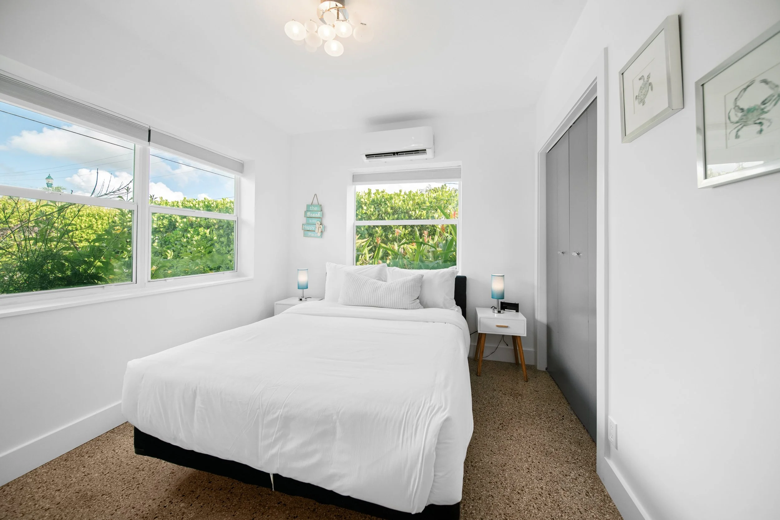 A bedroom with white walls and large windows showing green foliage outside. There is a white bed with pillows, a small white nightstand with a blue lamp, and framed art on the wall. An air conditioning unit is mounted above the windows.