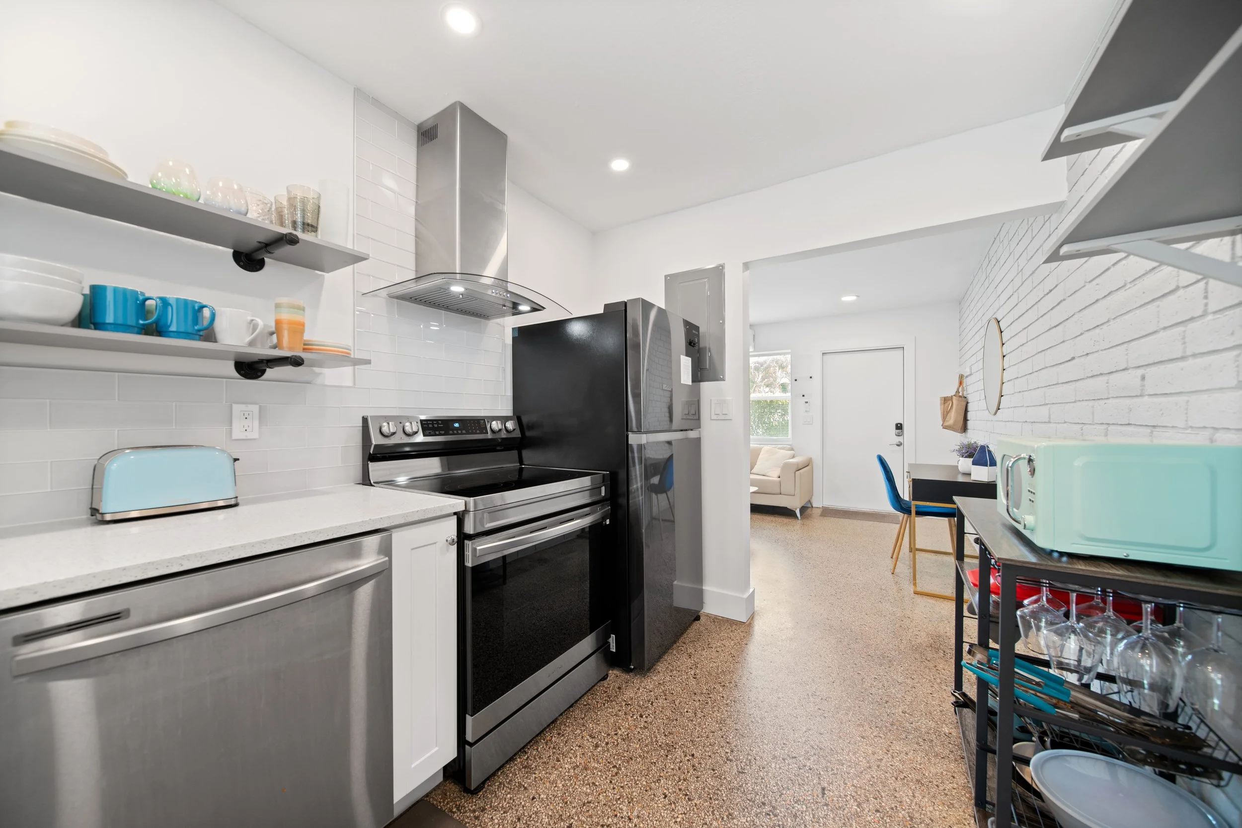 A modern kitchen with white cabinets, stainless steel appliances, open shelves with dishes, and a view into a living room with a sofa.