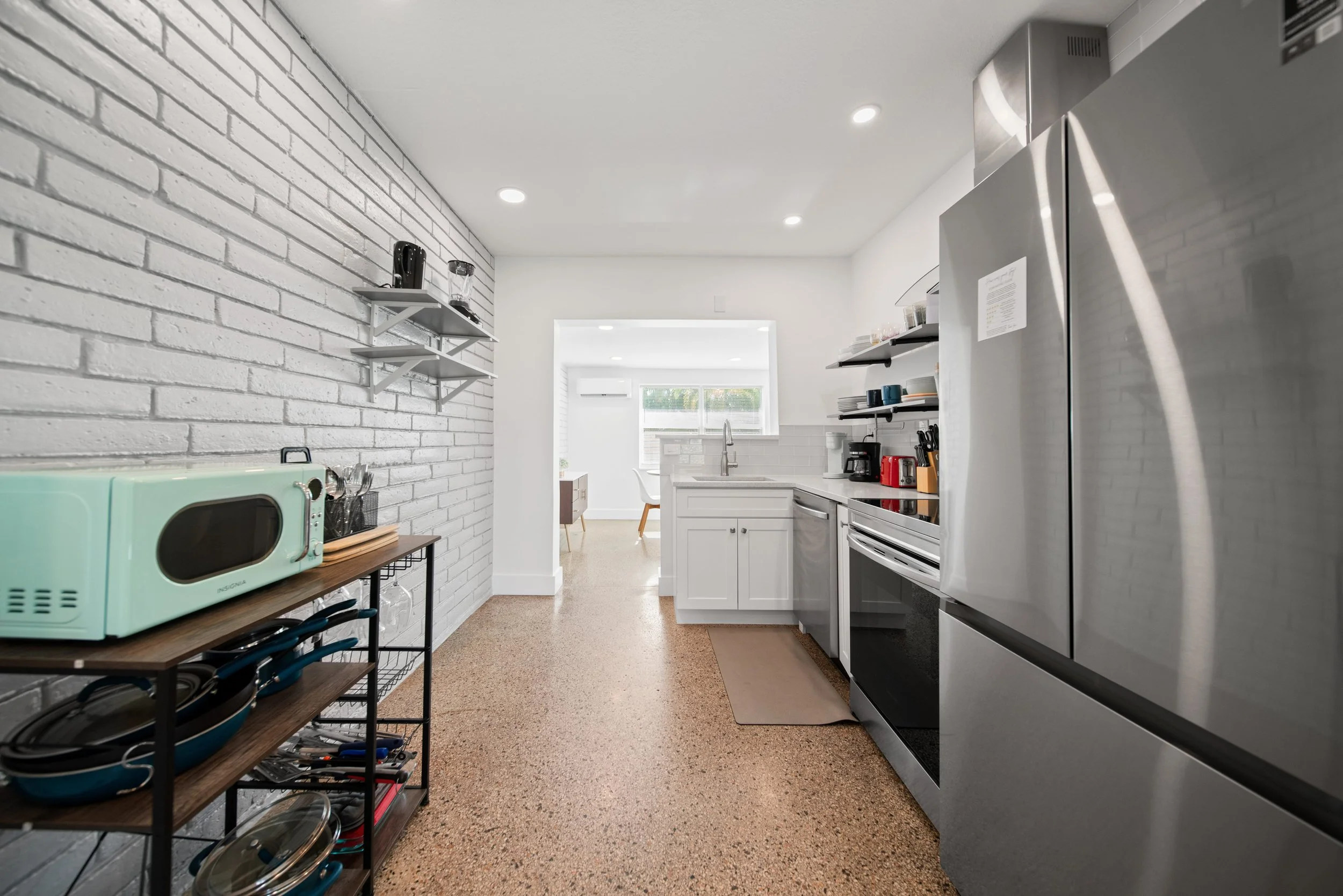 A modern kitchen with white brick wall on the left, white cabinets, a stainless steel refrigerator on the right, and black shelves with kitchenware. There is a microwave on a wooden table and a glimpse of a dining area with a window and chairs in the