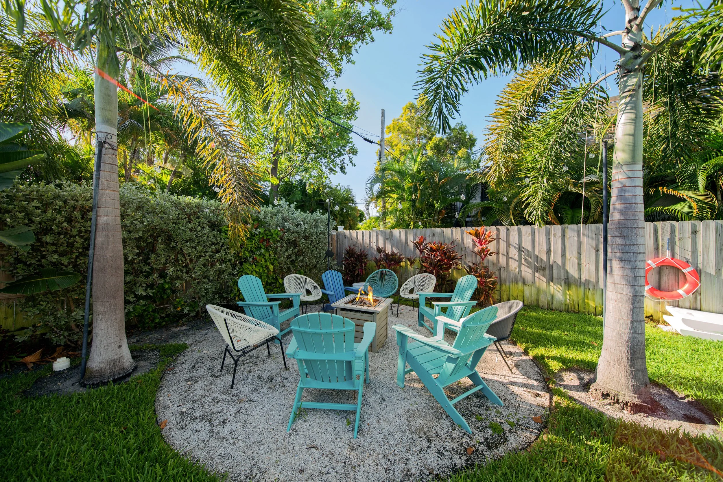 A backyard patio with turquoise, white, and black chairs arranged around a small fire pit, surrounded by lush tropical plants and tall palm trees under a blue sky.