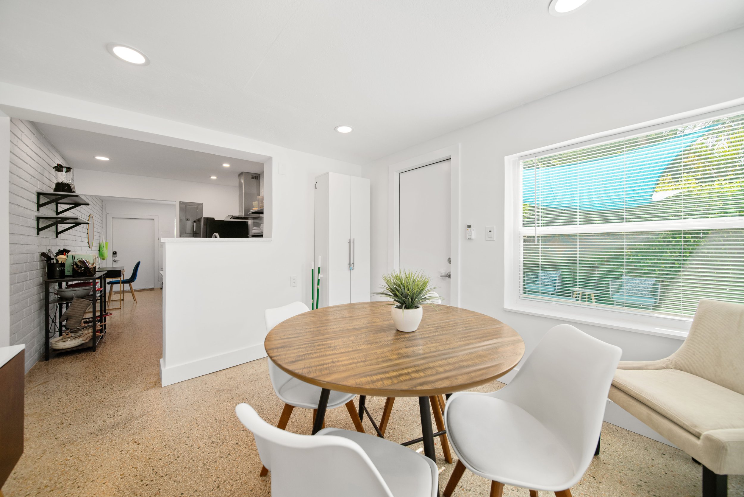 Bright, modern kitchen and dining area with a round wooden table, white chairs, large window with blinds, potted plant, and view of backyard.