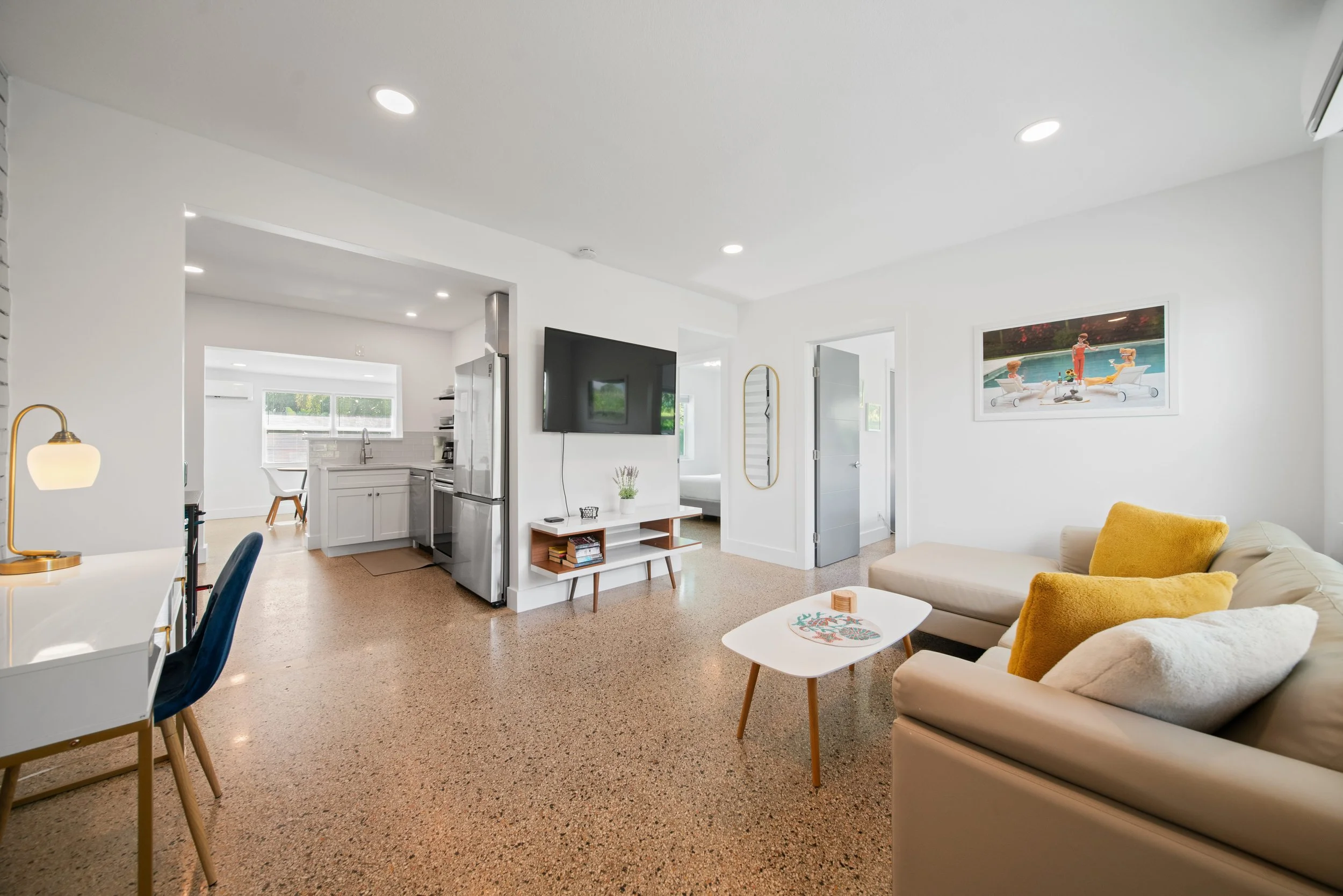 Living room with white walls and ceiling, a beige sofa with yellow pillows, a white coffee table, flat-screen TV on the wall, and artwork of people at the pool on the wall. The room is connected to a kitchen with white cabinets and refrigerator.