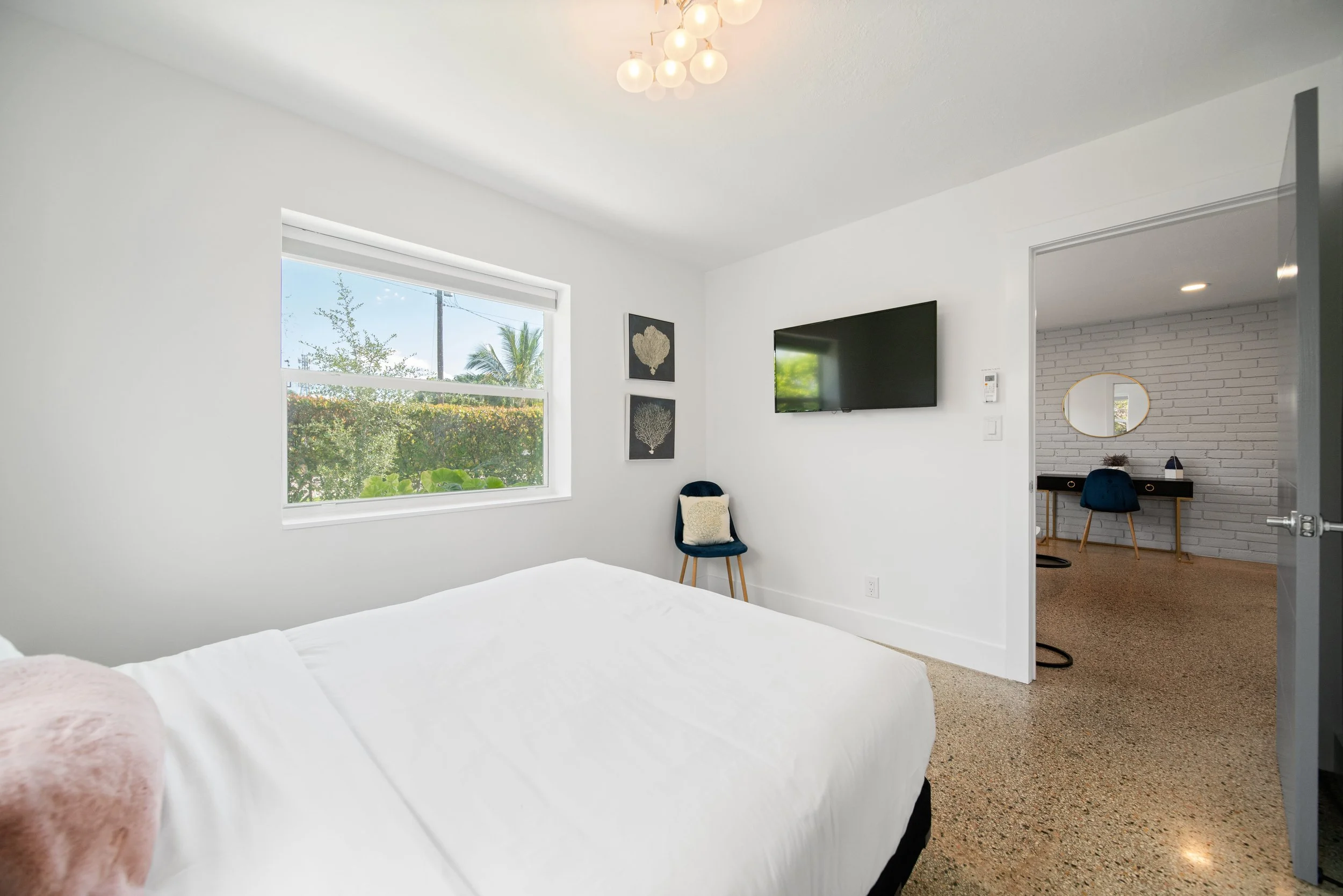 A bedroom with white walls, a window showing greenery outside, a wall-mounted flat-screen TV, a blue chair with a framed artwork above, and a partially visible white bed in the foreground.