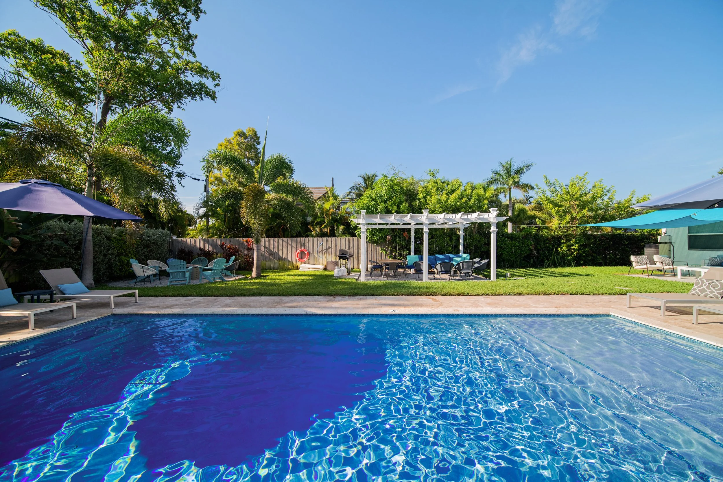 A backyard swimming pool with blue water, surrounded by lounge chairs, umbrellas, and lush greenery under a clear blue sky.
