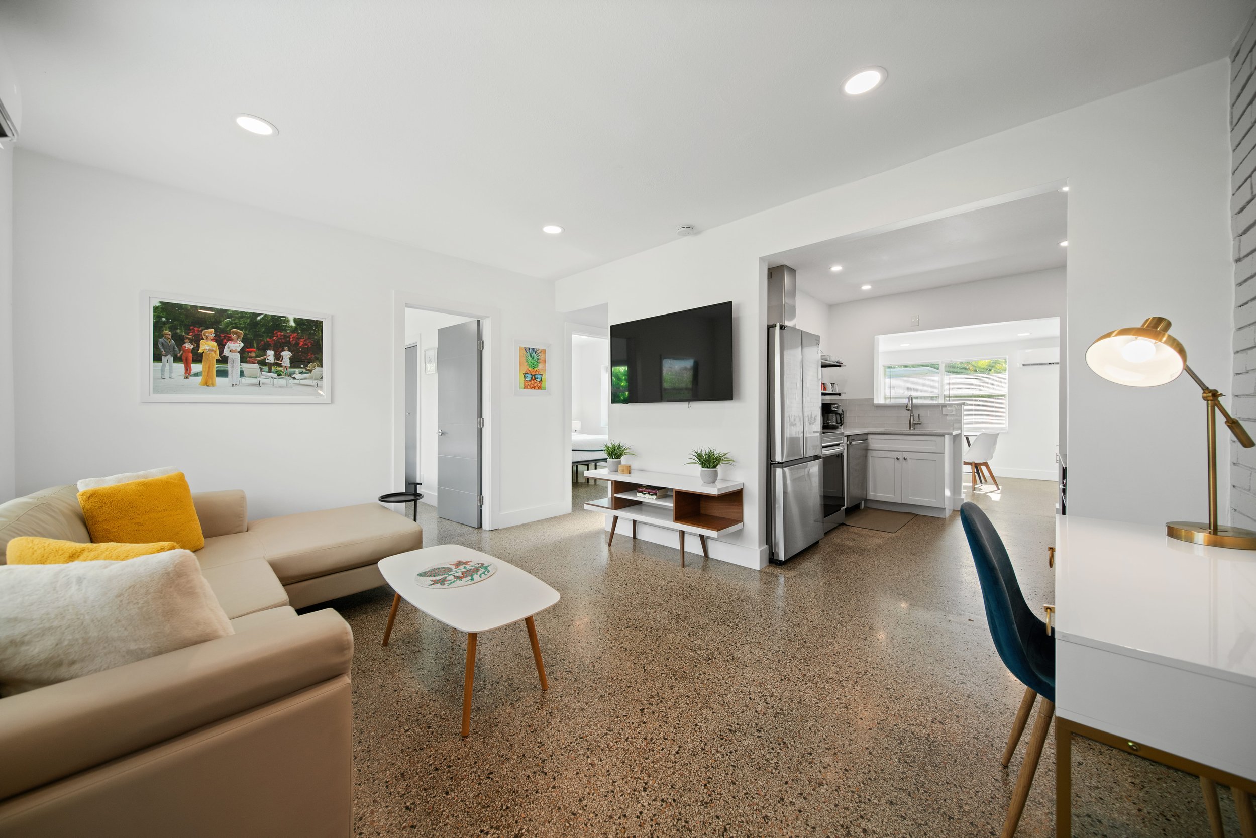 Living room with beige sofa, white coffee table, wall-mounted TV, kitchen with stainless steel appliances, and colorful artwork on white walls.