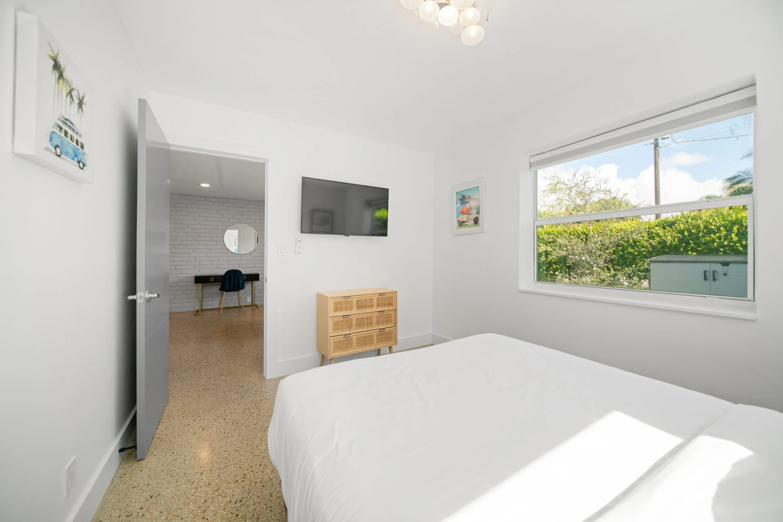 A bright, modern bedroom with a large window showing greenery outside. The room features a mounted flat-screen TV on a white wall, a wooden dresser, and colorful wall art including a framed beach scene and a canvas with a van and palm trees. Part of 