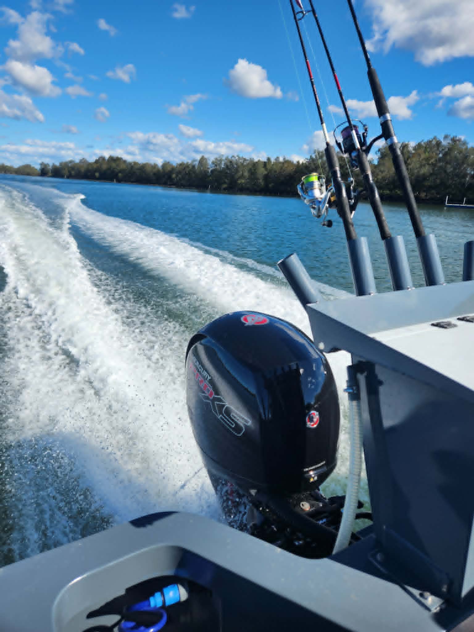 View from a boat showing two fishing poles attached to the boat, with a waterway and a treeline in the background under a mostly clear blue sky with some white clouds.
