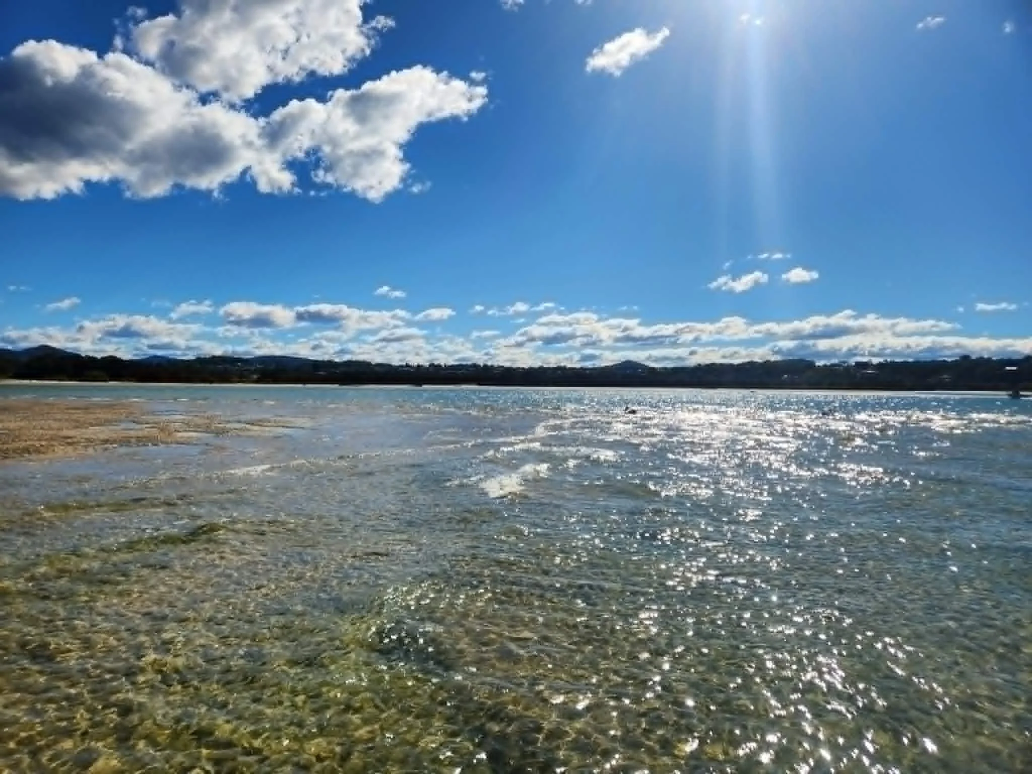 A landscape scene of a body of water with a sandy shoreline, distant hills, and a bright blue sky with some white clouds and sun rays.