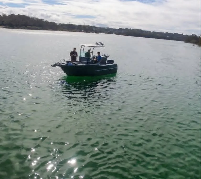A small boat on a body of water with three people on board, one standing and two seated. The boat has fishing equipment on it, and the water is reflecting sunlight, with a distant shoreline visible under a cloudy sky.