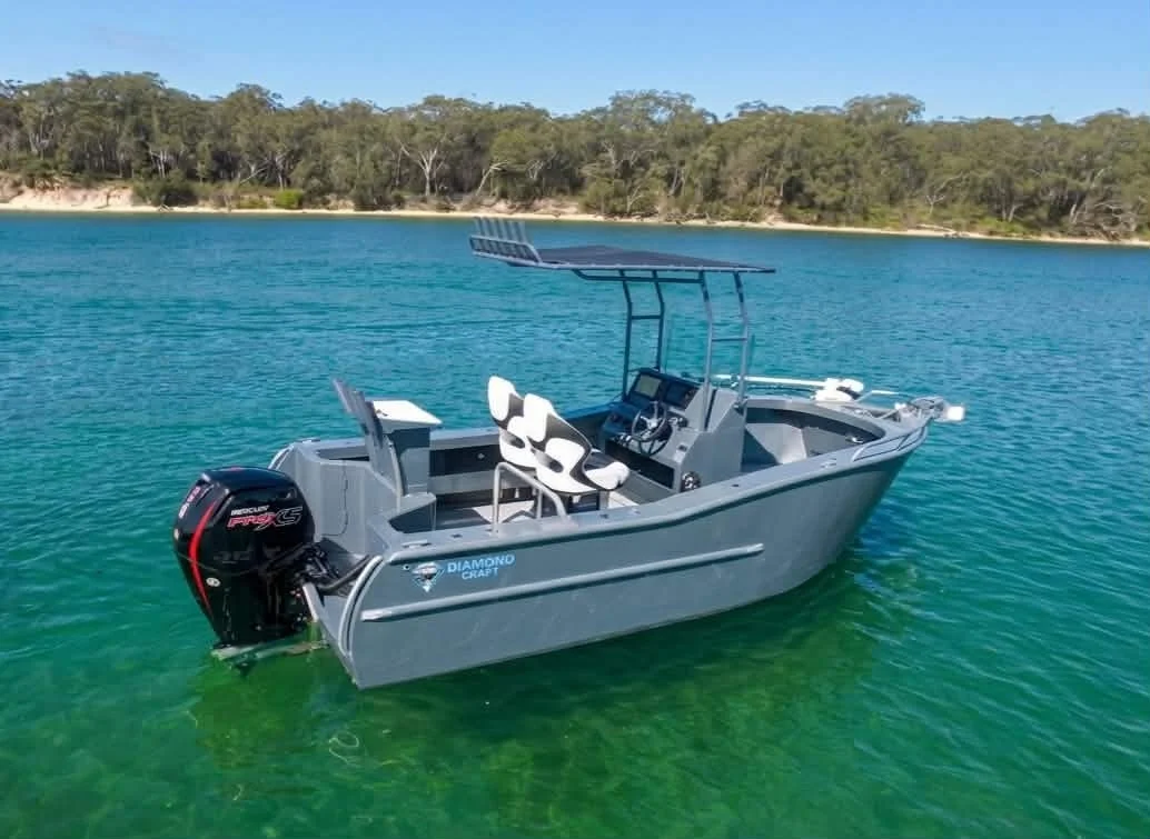 A gray boat with white seats floating on clear green water near a forested shoreline under a blue sky.
