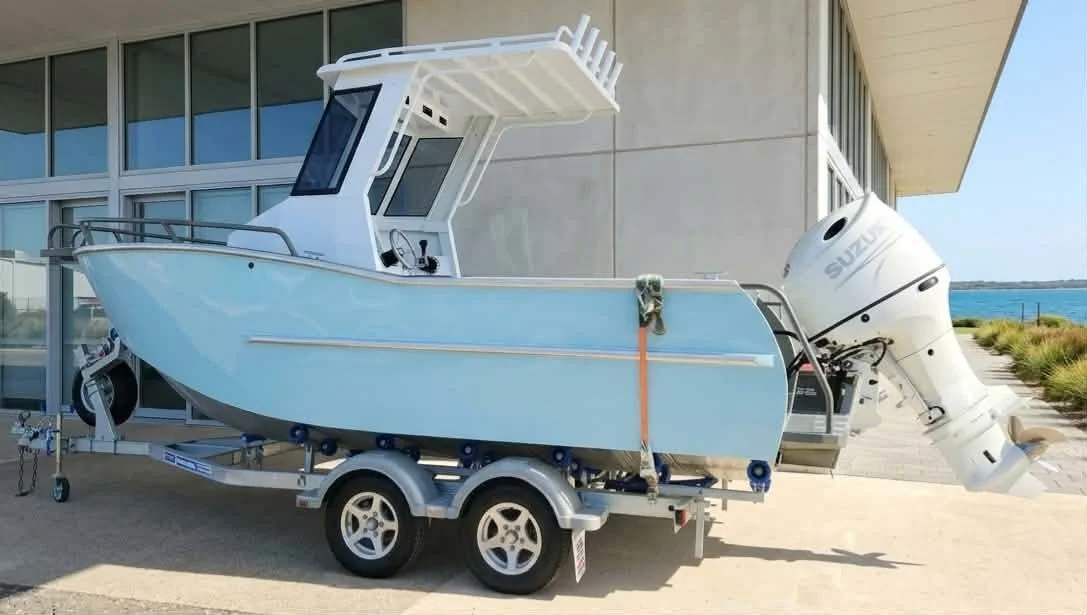 A small motorboat on a trailer with a large outboard engine attached, parked in front of a building near the water.