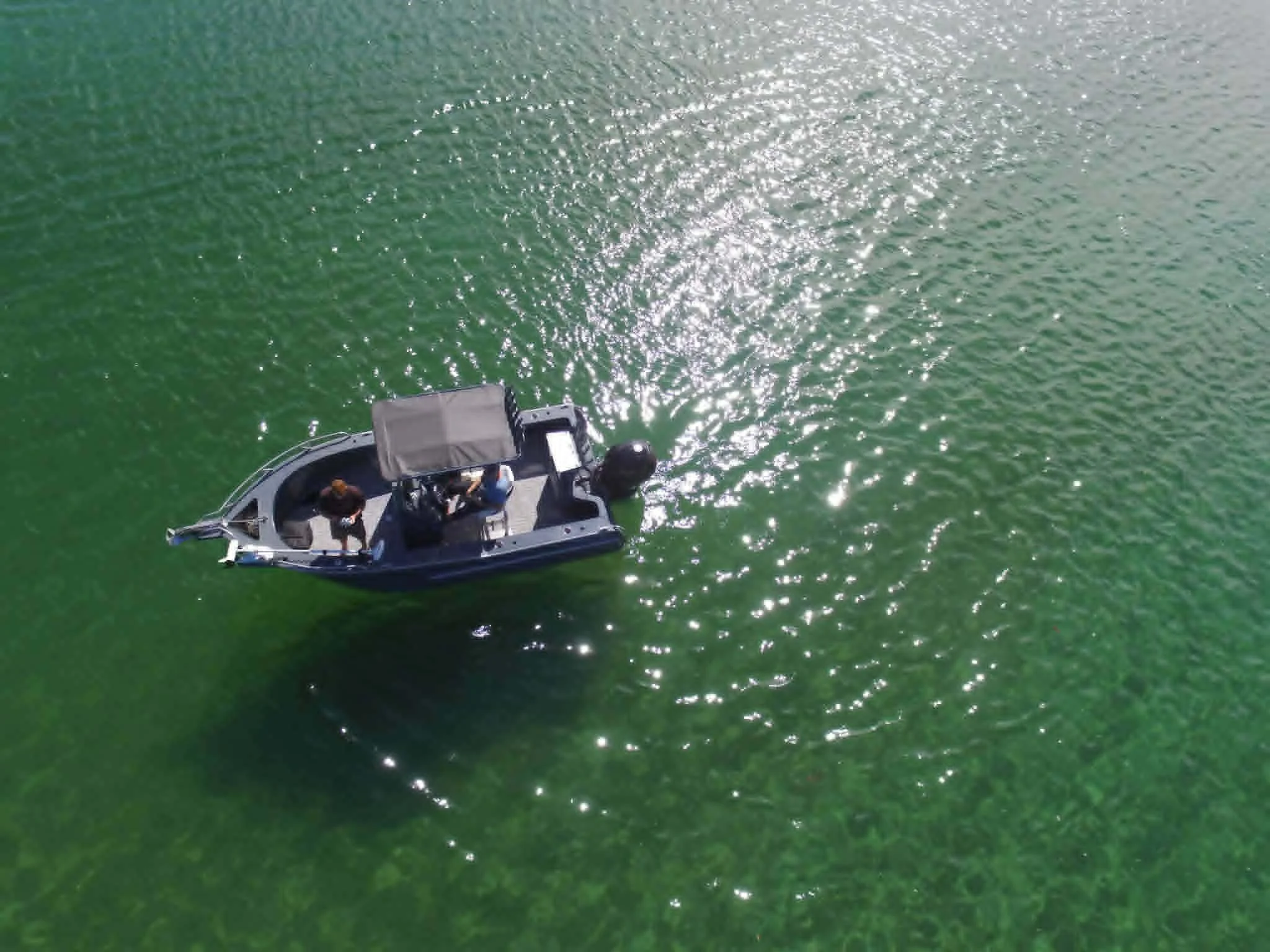A boat on green water with three people on board, some sitting and some standing, with the water sparkling under sunlight.