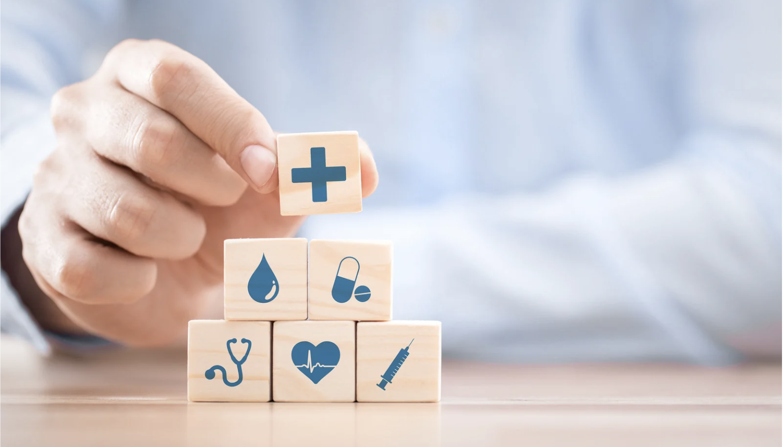 A person arranging wooden blocks with medical symbols, including a plus sign, drop of liquid, pills, stethoscope, heartbeat, and syringe.