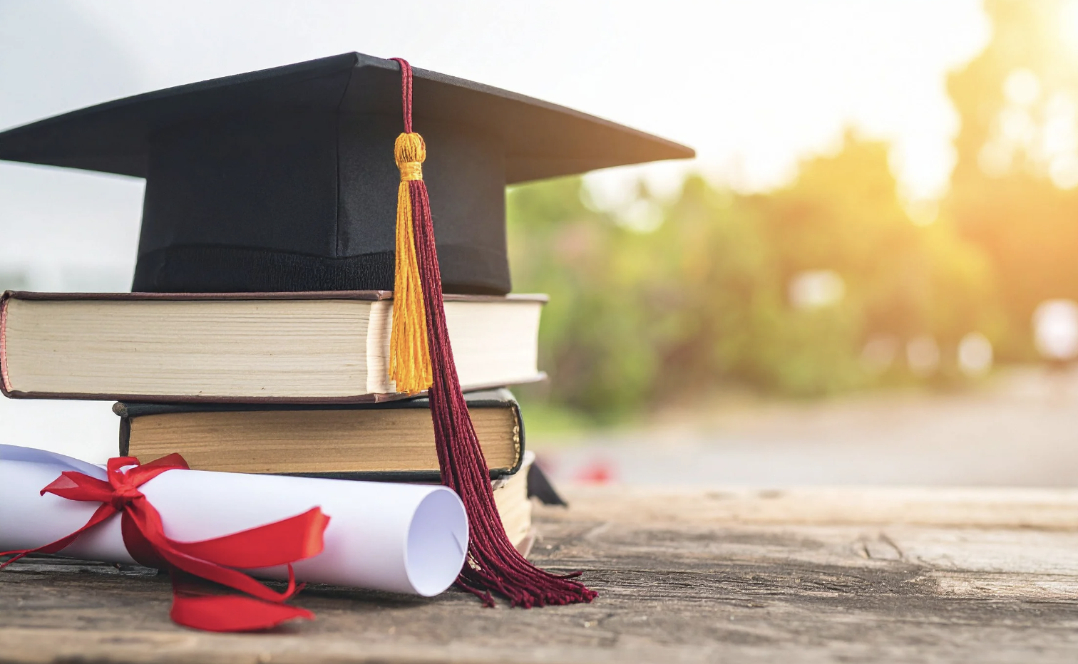A black graduation cap with a red and yellow tassel, placed on top of a stack of three books, with a rolled diploma tied with a red ribbon in front of the books, on a wooden surface outdoors during sunset.