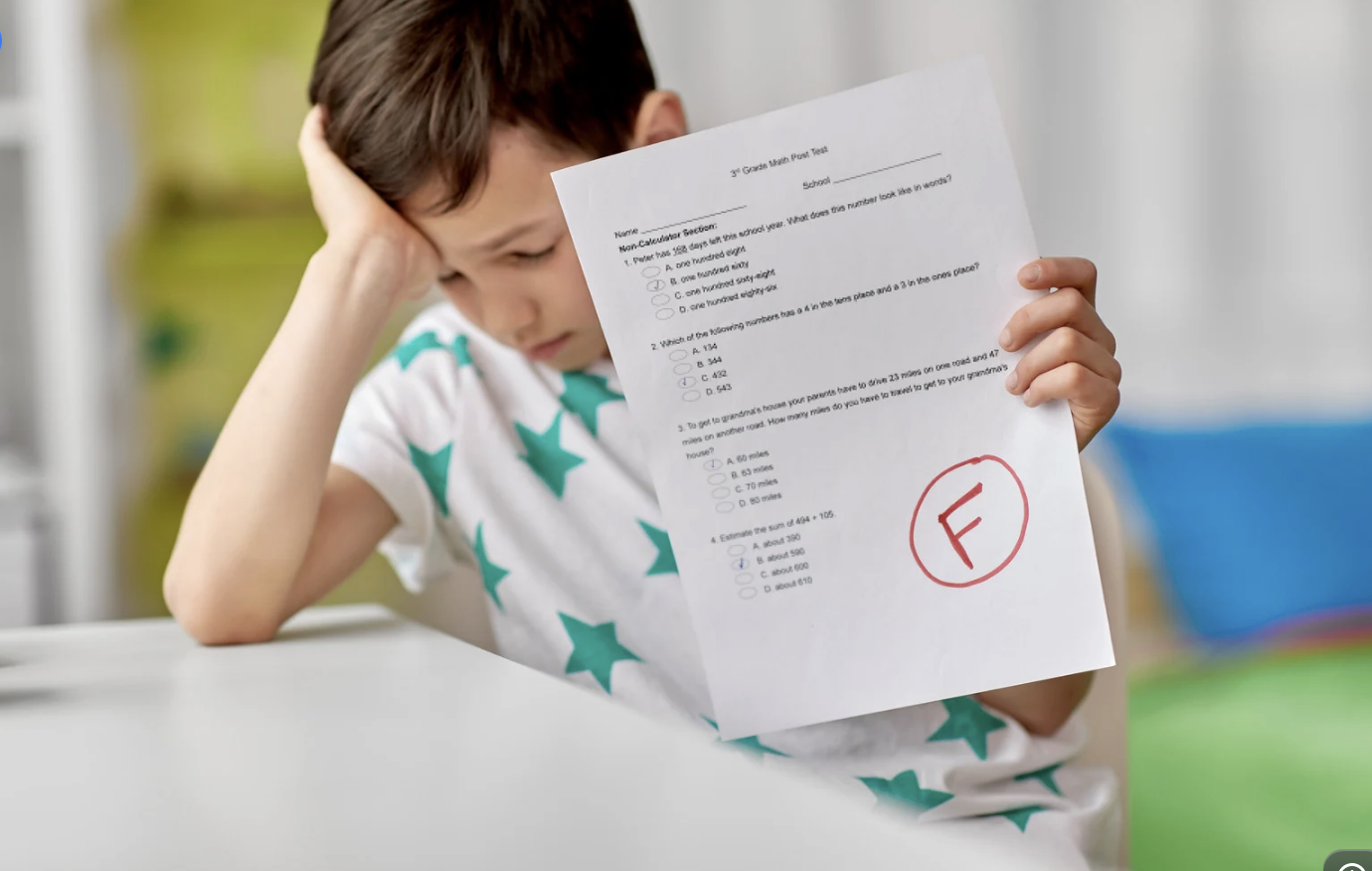 A young boy with brown hair, wearing a white shirt with teal stars, sitting at a white table, holding a test paper with a red circle and a large 'F' grade, looking distressed.