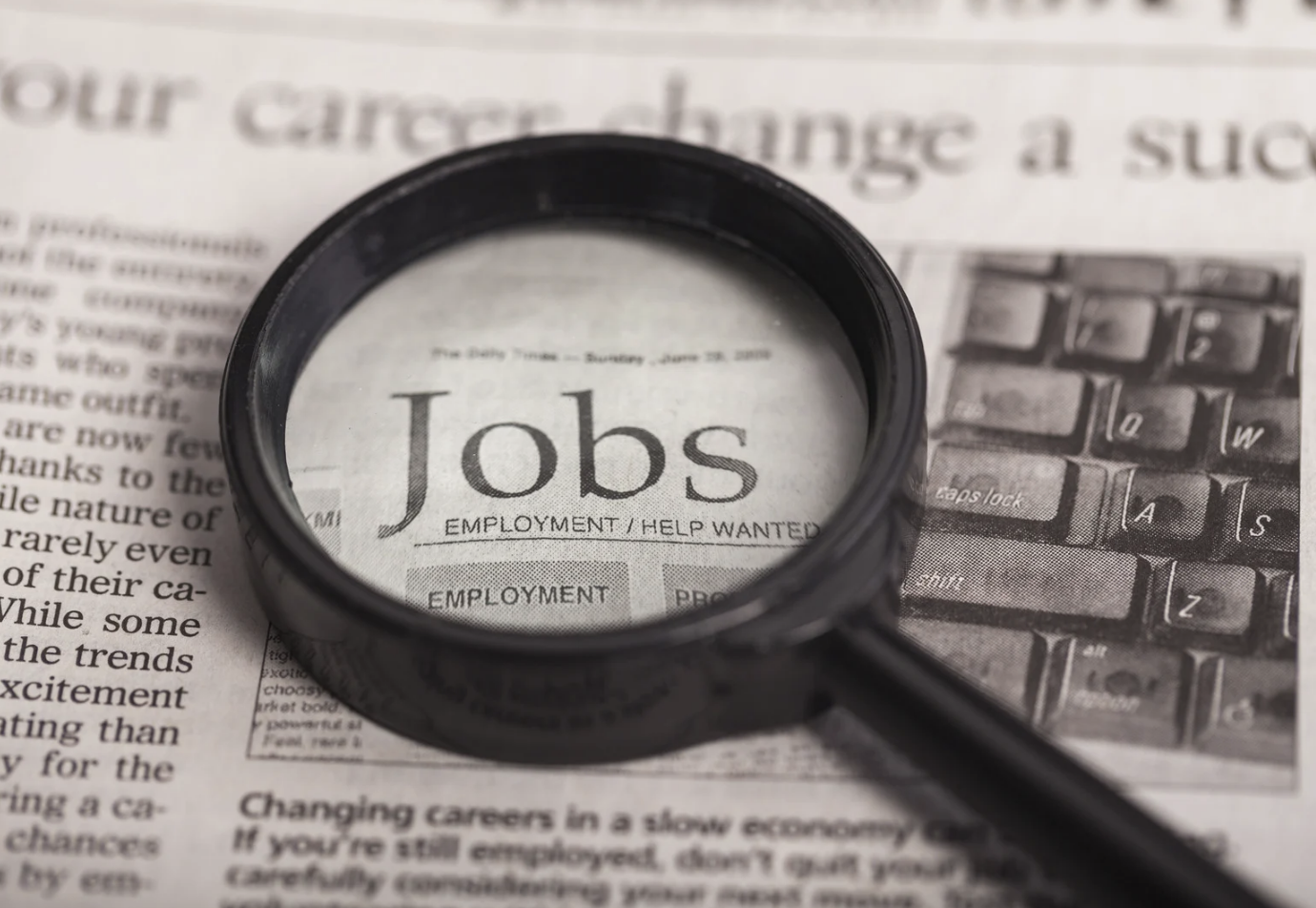 A magnifying glass focusing on a newspaper with a headline that reads 'Jobs' and section 'Employment/Help Wanted', alongside a computer keyboard.