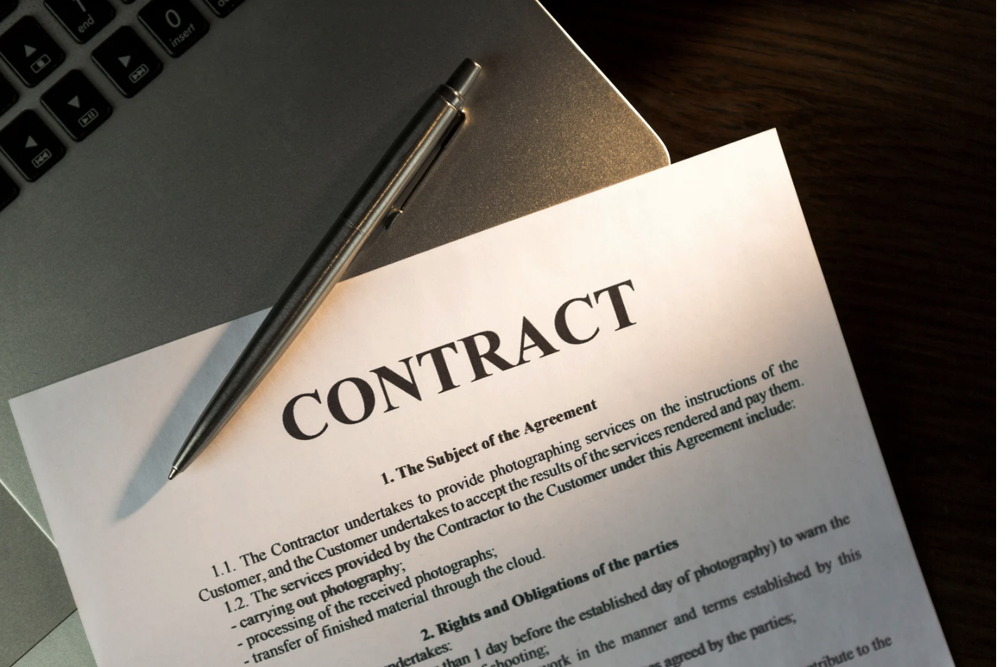 Contract paper with the title 'CONTRACT' and a silver pen on top, placed on a silver laptop and dark wooden surface.
