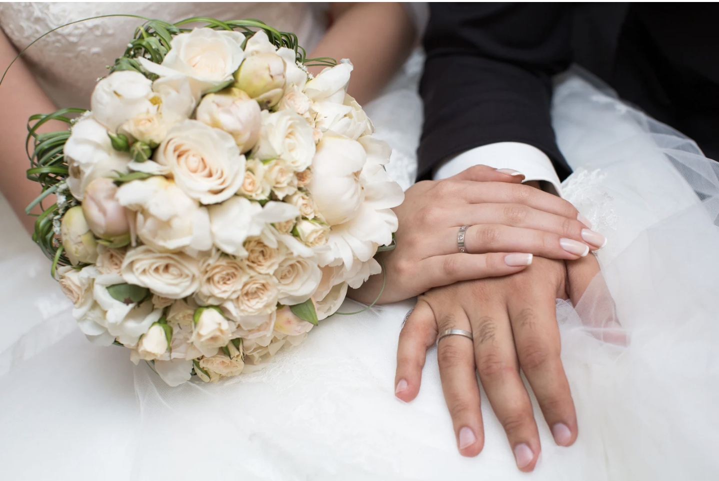 A bride and groom holding hands, with wedding rings, resting on a white wedding dress, beside a bouquet of white and blush roses and peonies.