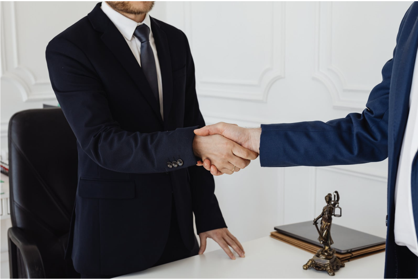 Two men in suits shaking hands in an office, with a desk, a statue, and folders in the background.