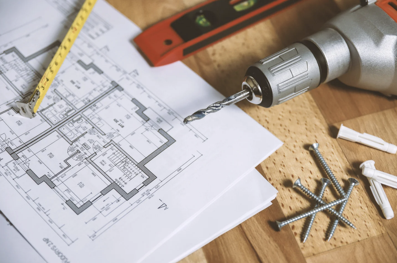 Blueprints of a building, a yellow measuring tape, a corded power drill with a drill bit, several screws, and wall anchors on a wooden surface.