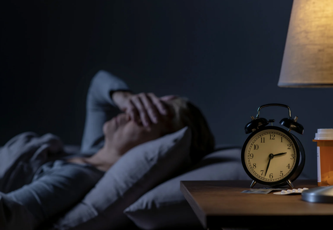 A person lying in bed holding their head in distress, with an alarm clock showing 3:12 on a bedside table, in a dimly lit room, suggesting the person has difficulty sleeping or is experiencing insomnia.