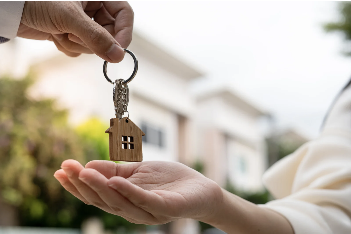 Person holding out a hand to receive a house-shaped keychain being handed over by another person outdoors.