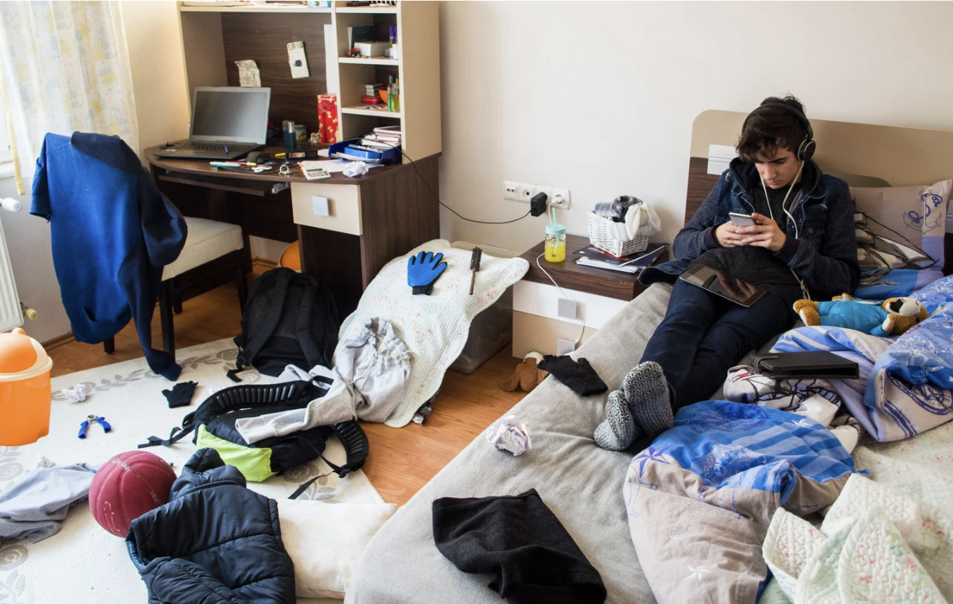 A teenage boy wearing headphones sitting on a cluttered bed using a smartphone in a messy bedroom with clothes, shoes, and personal items scattered around.