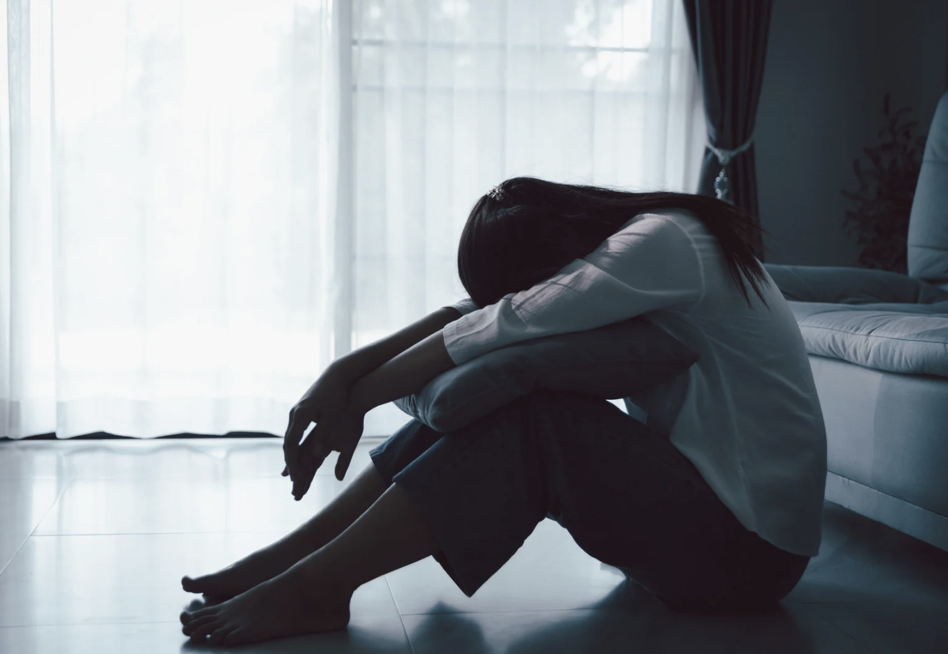 A woman sitting on the floor of a bedroom with her head resting on her knees, appearing sad or contemplative, with sunlight coming through a window in the background.