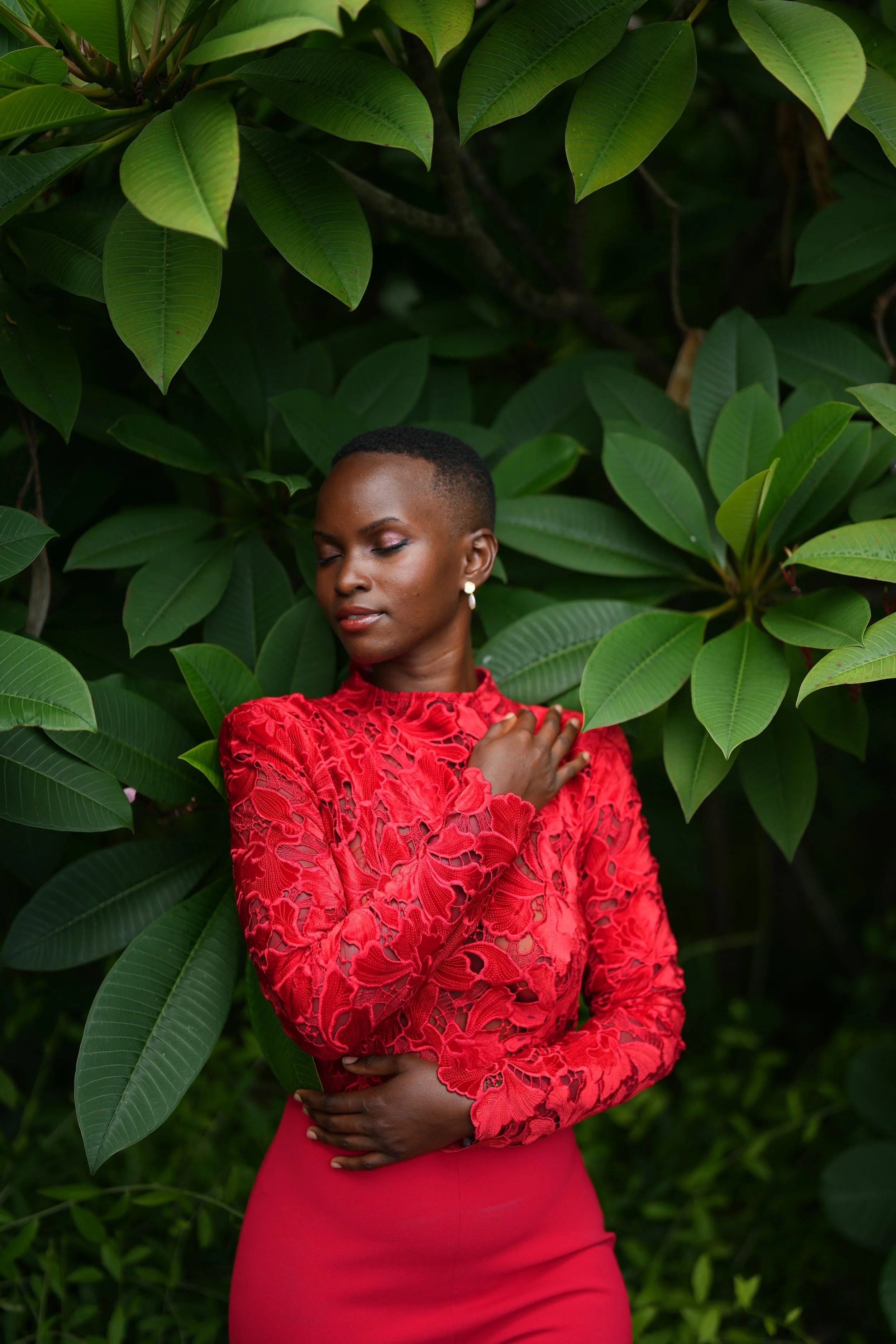 A woman wearing a red lace top and matching skirt standing among green leaves with her eyes closed, gently touching her chest.