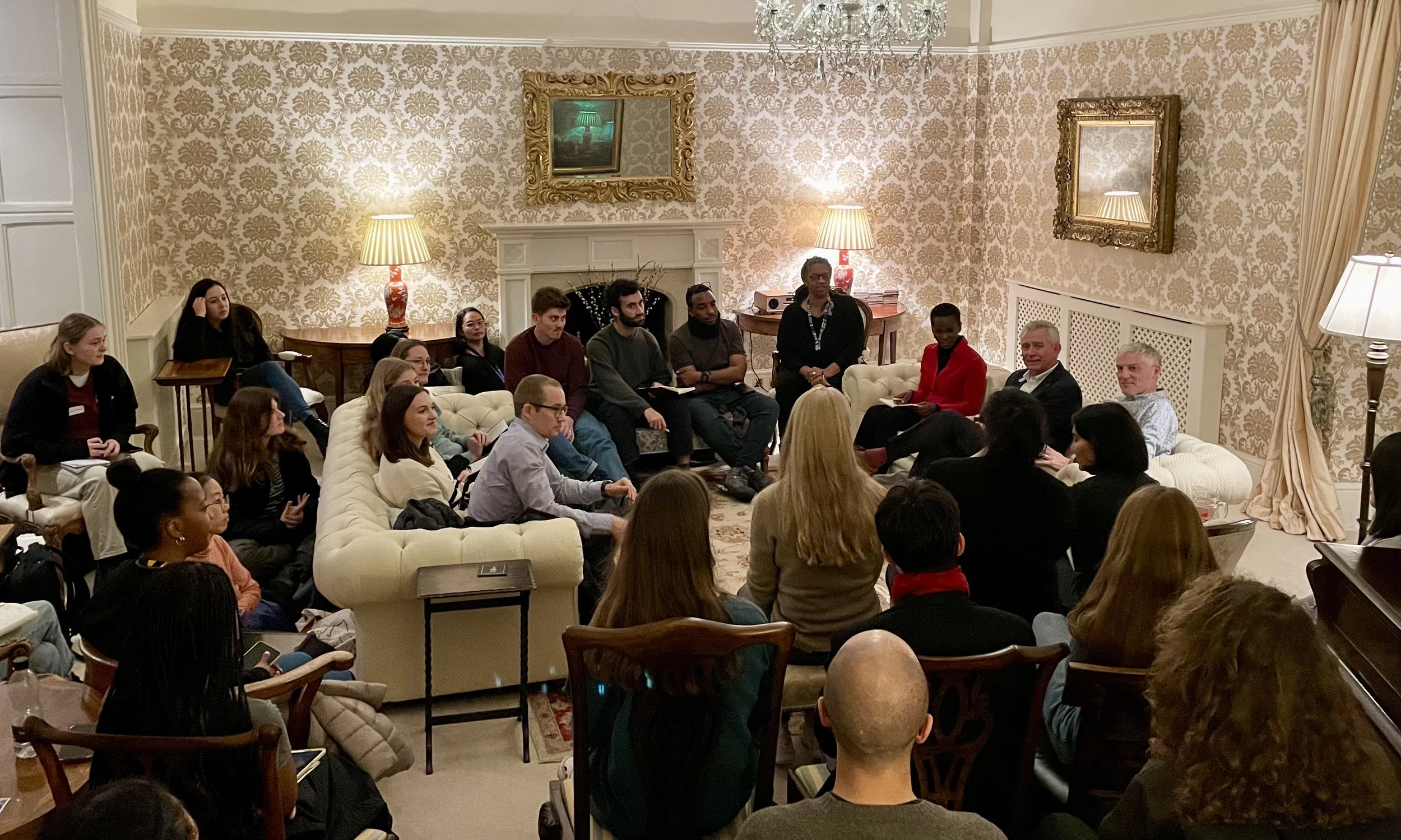 Group of people seated and standing in a formal living room with ornate wallpaper, chandeliers, and decorative mirrors, attending a meeting or discussion.