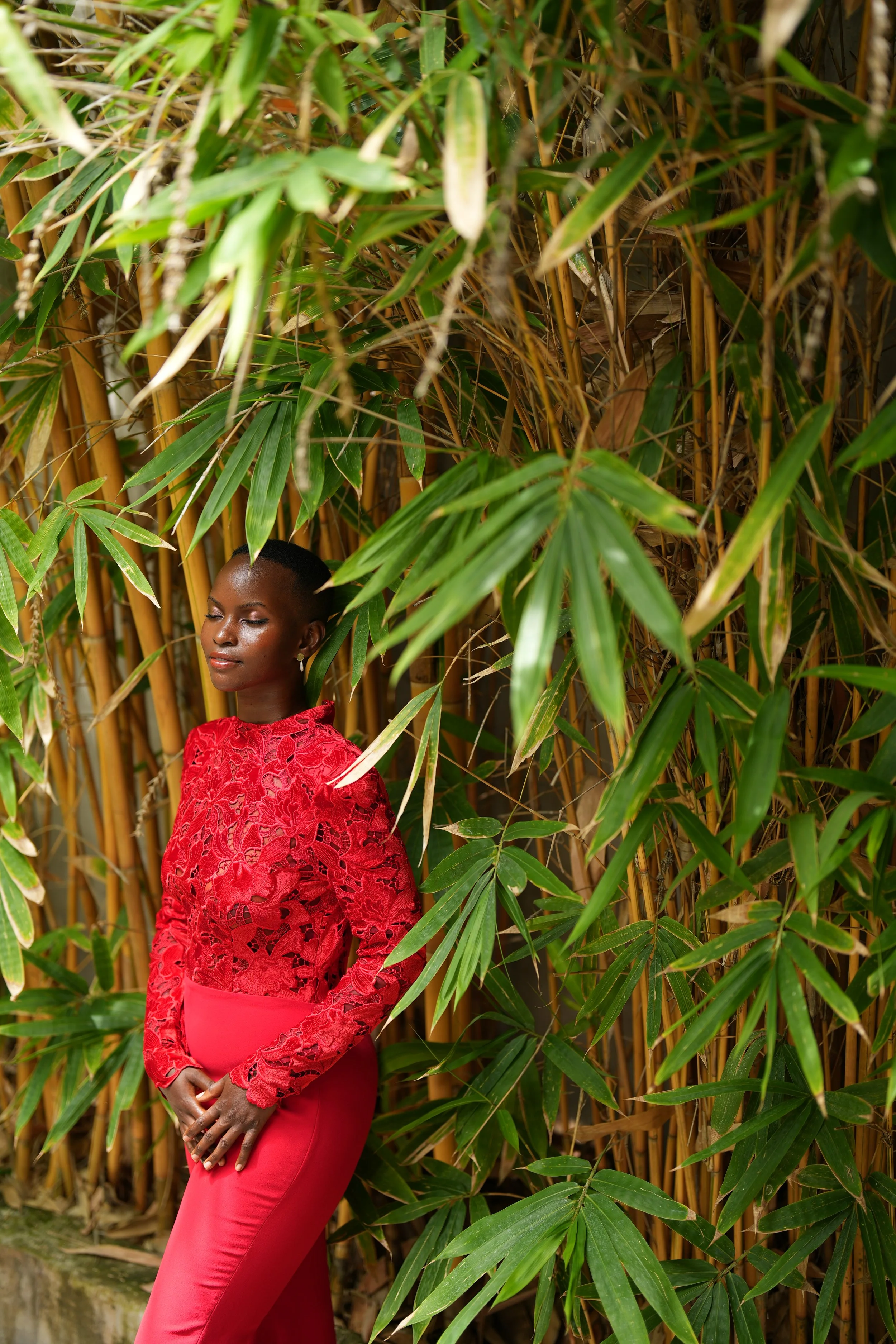 A woman in a red lace dress standing in front of tall bamboo plants with green leaves and stalks, her eyes closed and hands clasped in front of her.