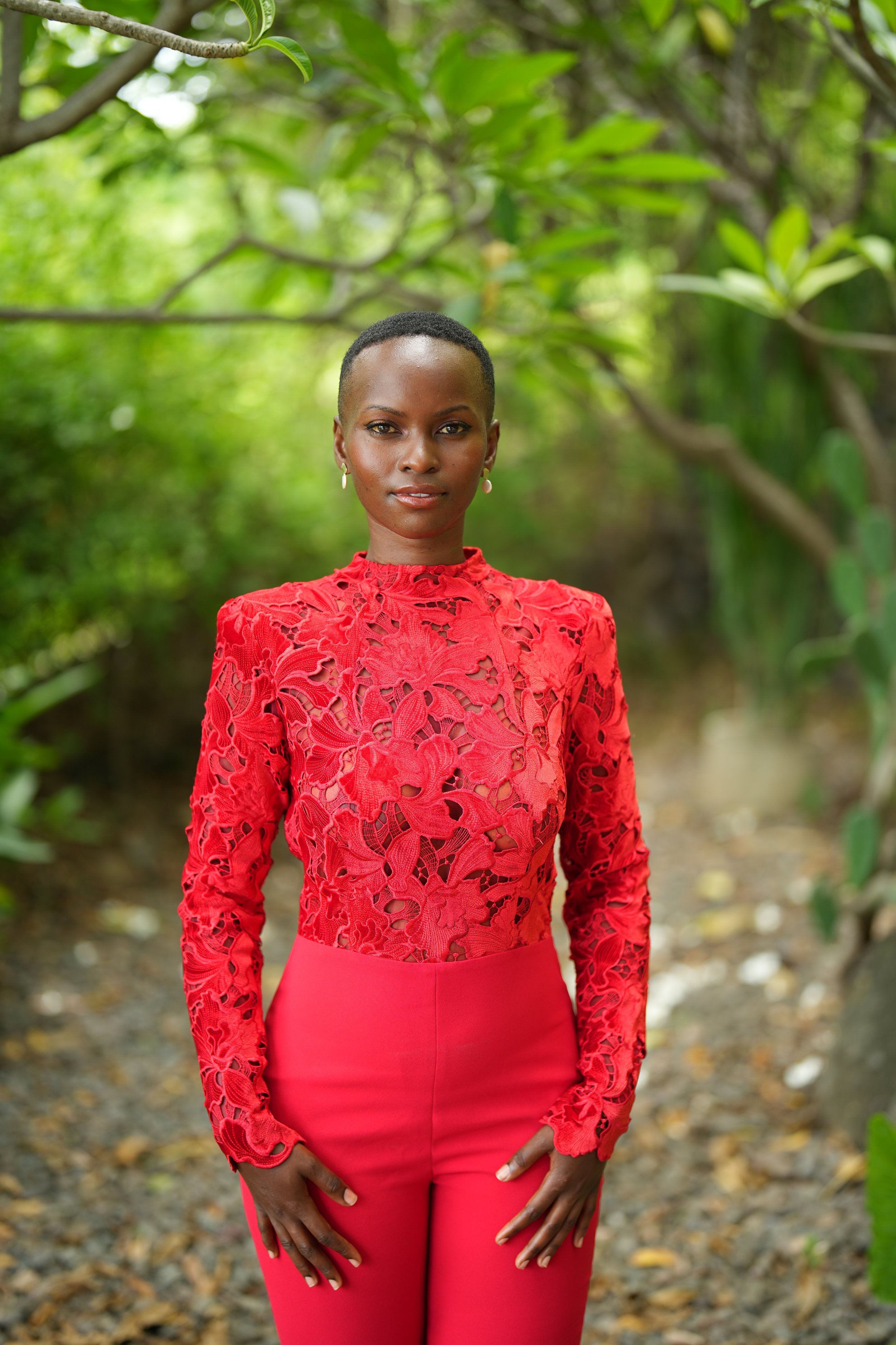 A woman standing outdoors among green trees, wearing a bright red lace top and matching red pants, looking at the camera with a confident expression.