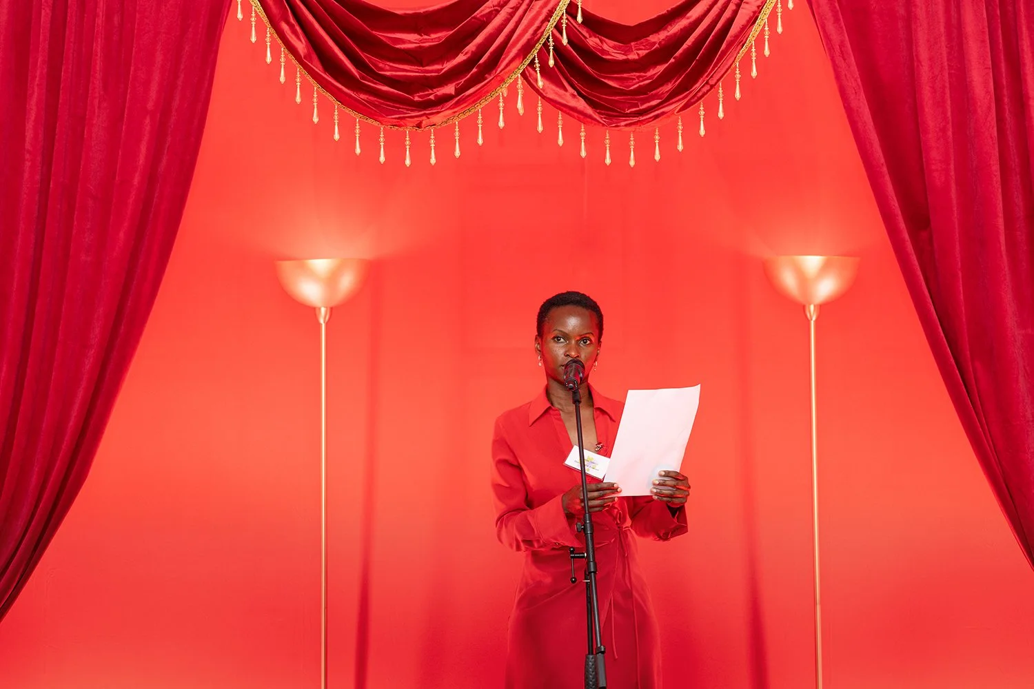 A woman dressed in red, standing in front of a microphone, reading from a sheet of paper, with a red curtain backdrop and two tall lamps on either side.