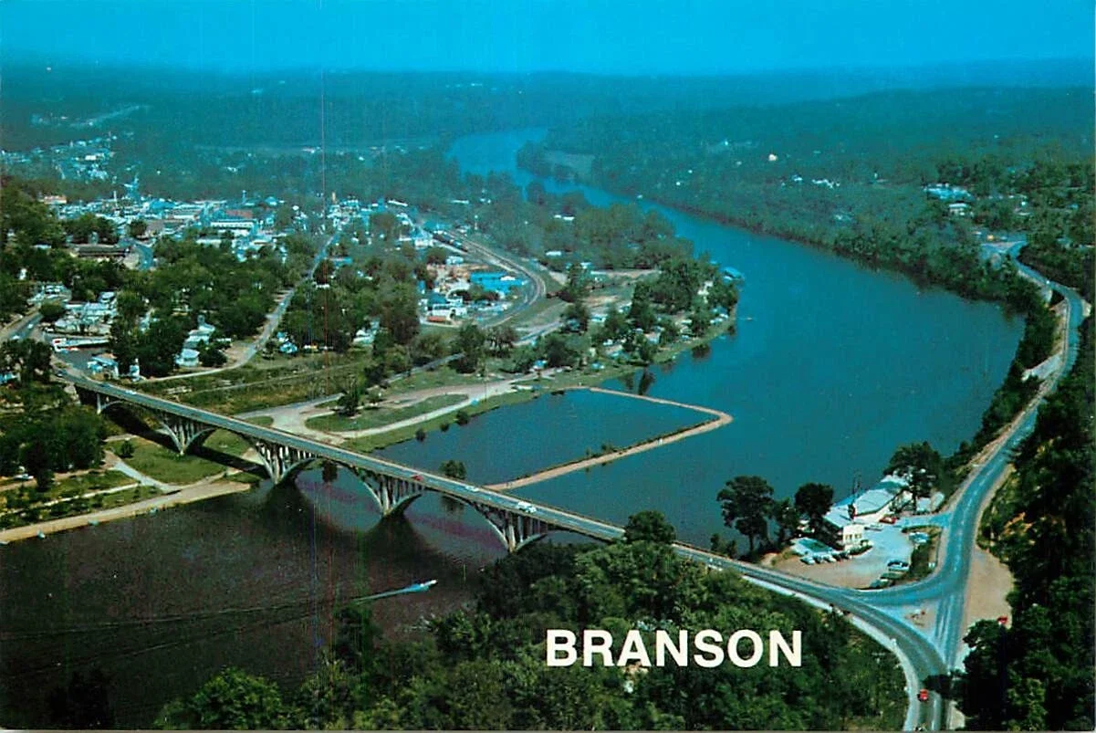 Aerial view of Branson, Missouri, showing a river, a bridge, and surrounding town with greenery and roads.