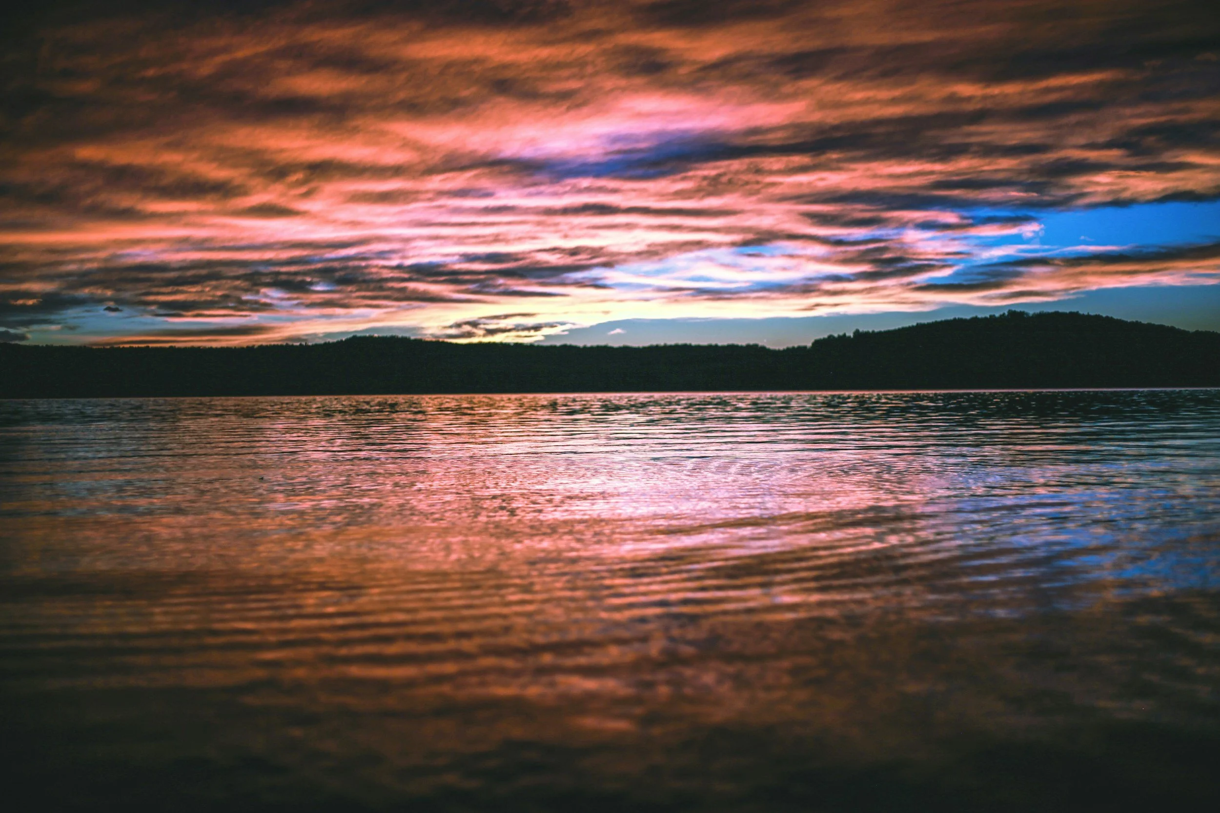 A colorful sunset over a body of water with dark hills in the distance and clouds illuminated with pink, purple, and blue hues.